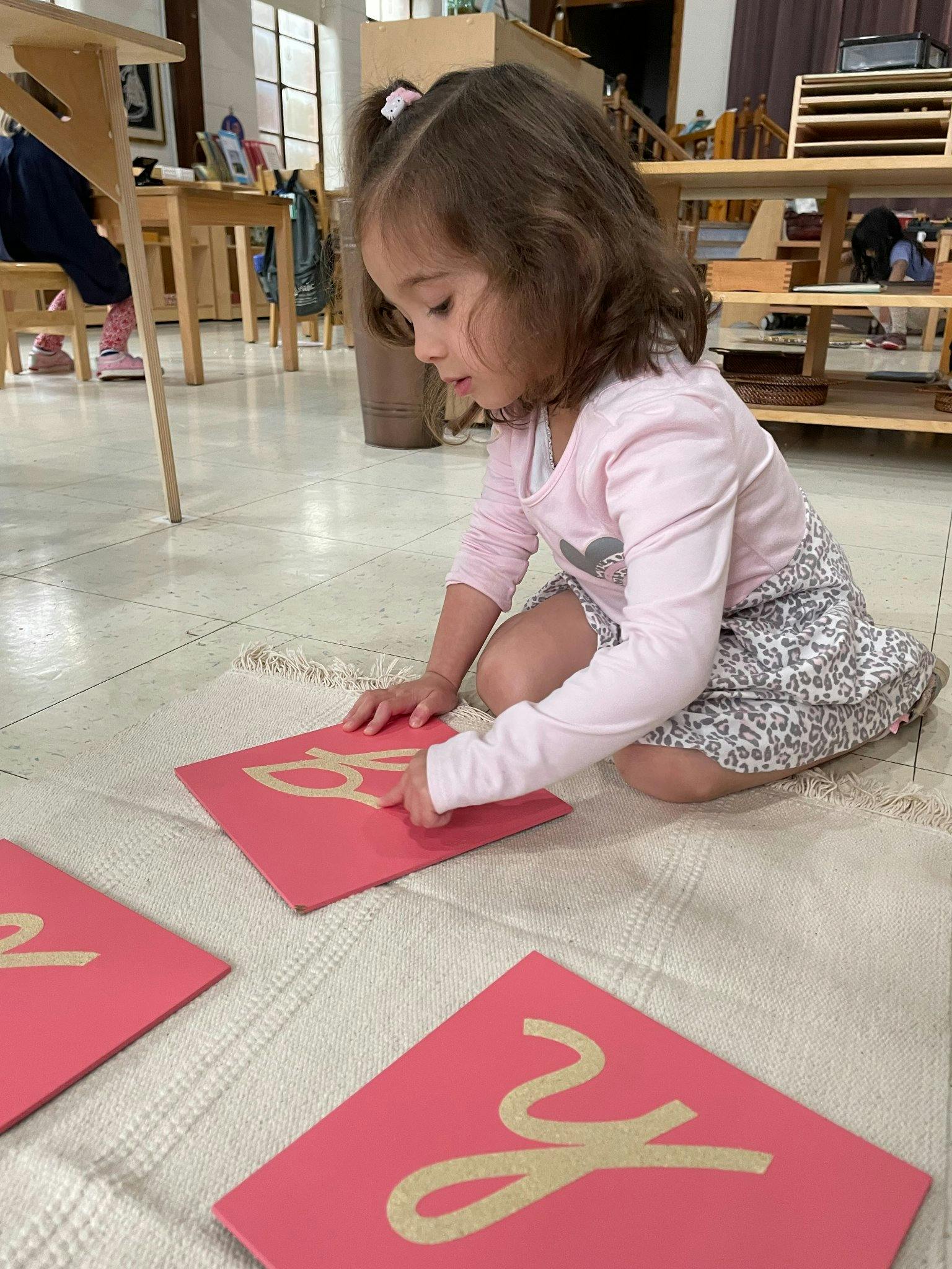 Child kneeling on a rug, tracing a letter on a red card