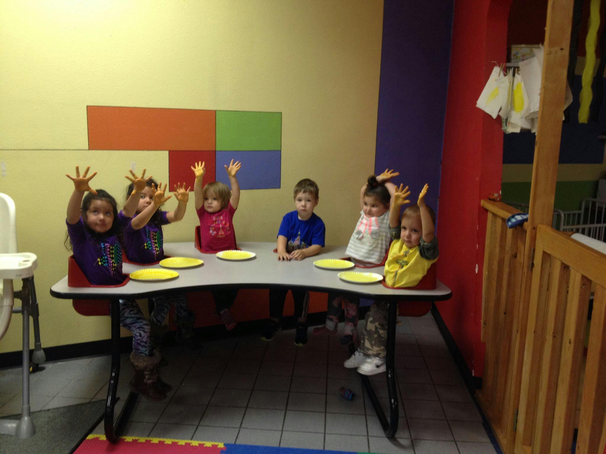 Children sitting at a table with yellow plates, some raising their hands