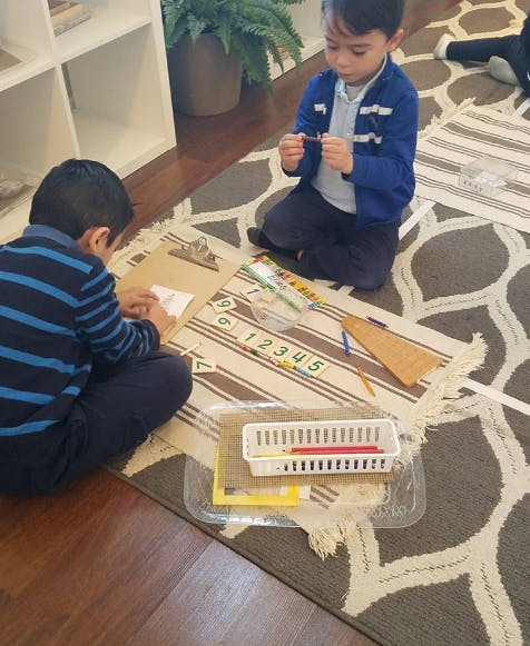 Two children sitting on a rug, one writing on paper, the other holding a crayon