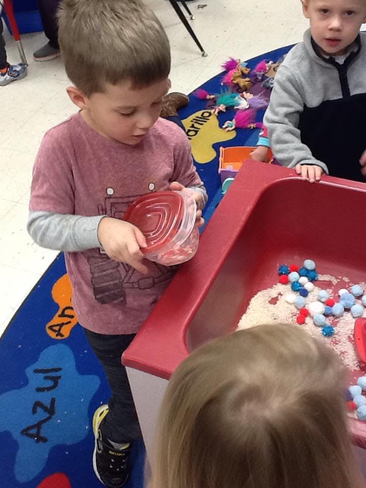 Child holding a container over a sensory bin with rice and pom-poms