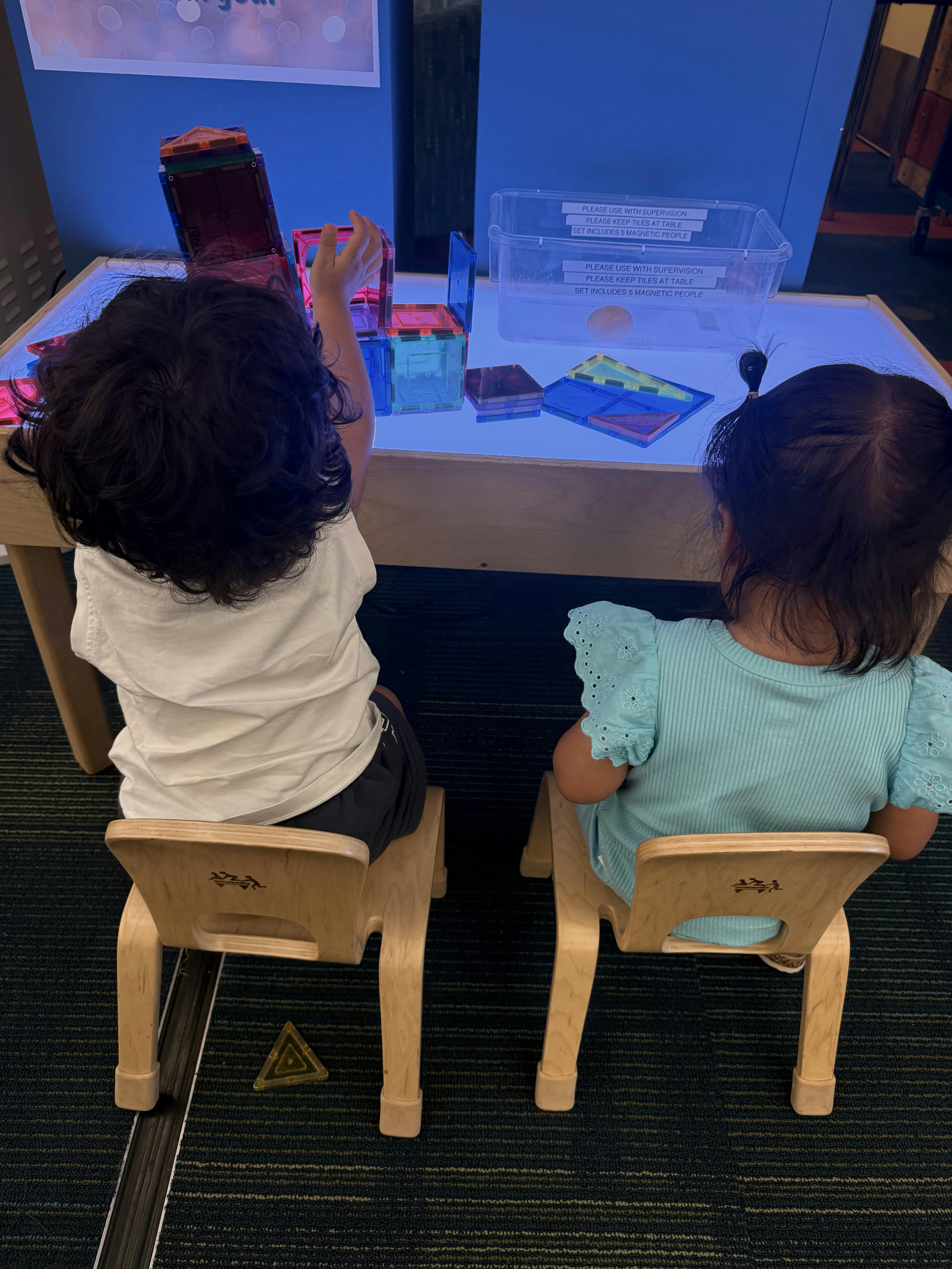 Two children sitting at a light table playing with colorful magnetic tiles