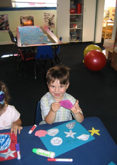 Child holding a purple cutout shape at a table with craft supplies
