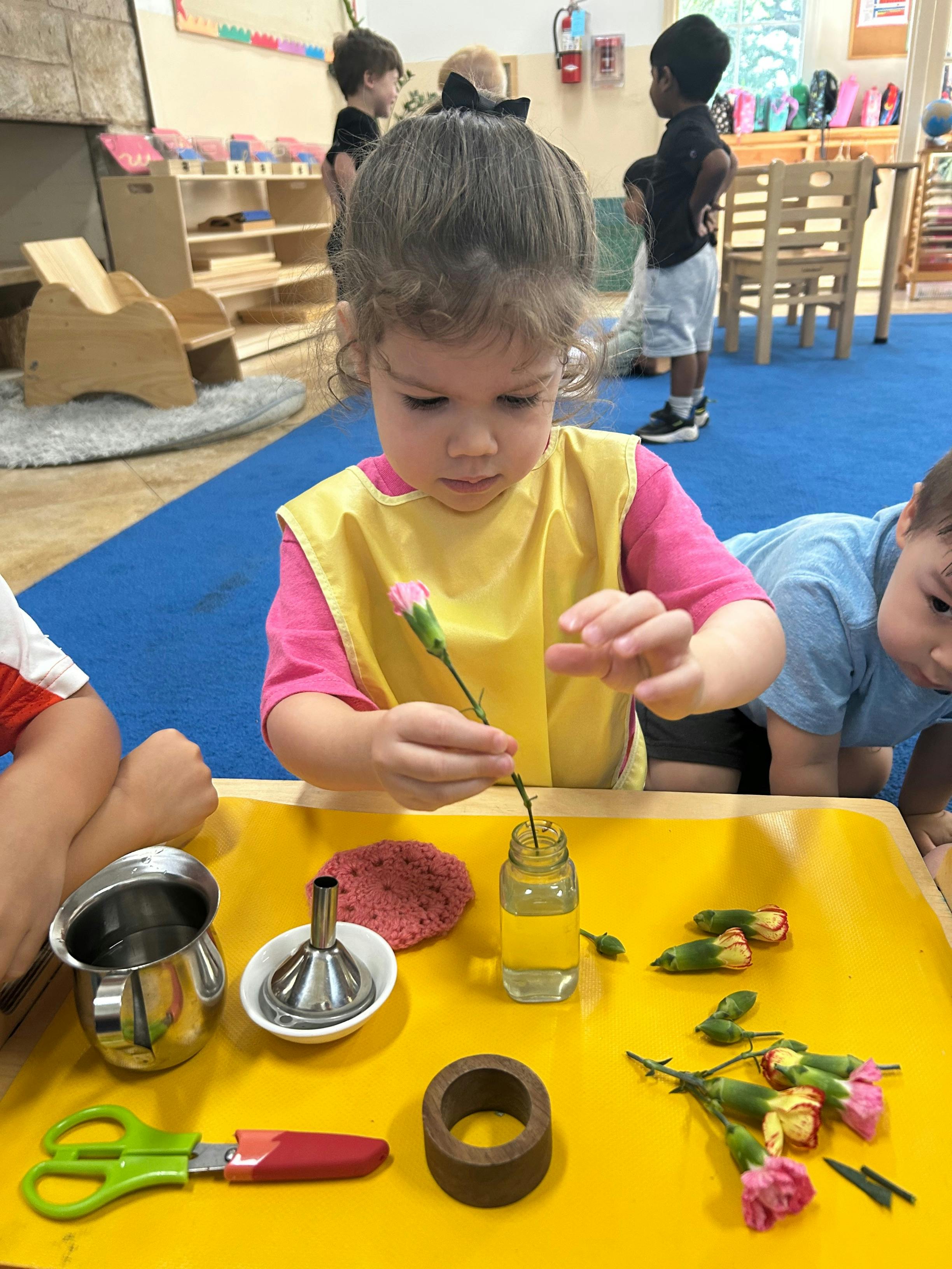 Child in a yellow bib putting a flower into a small jar