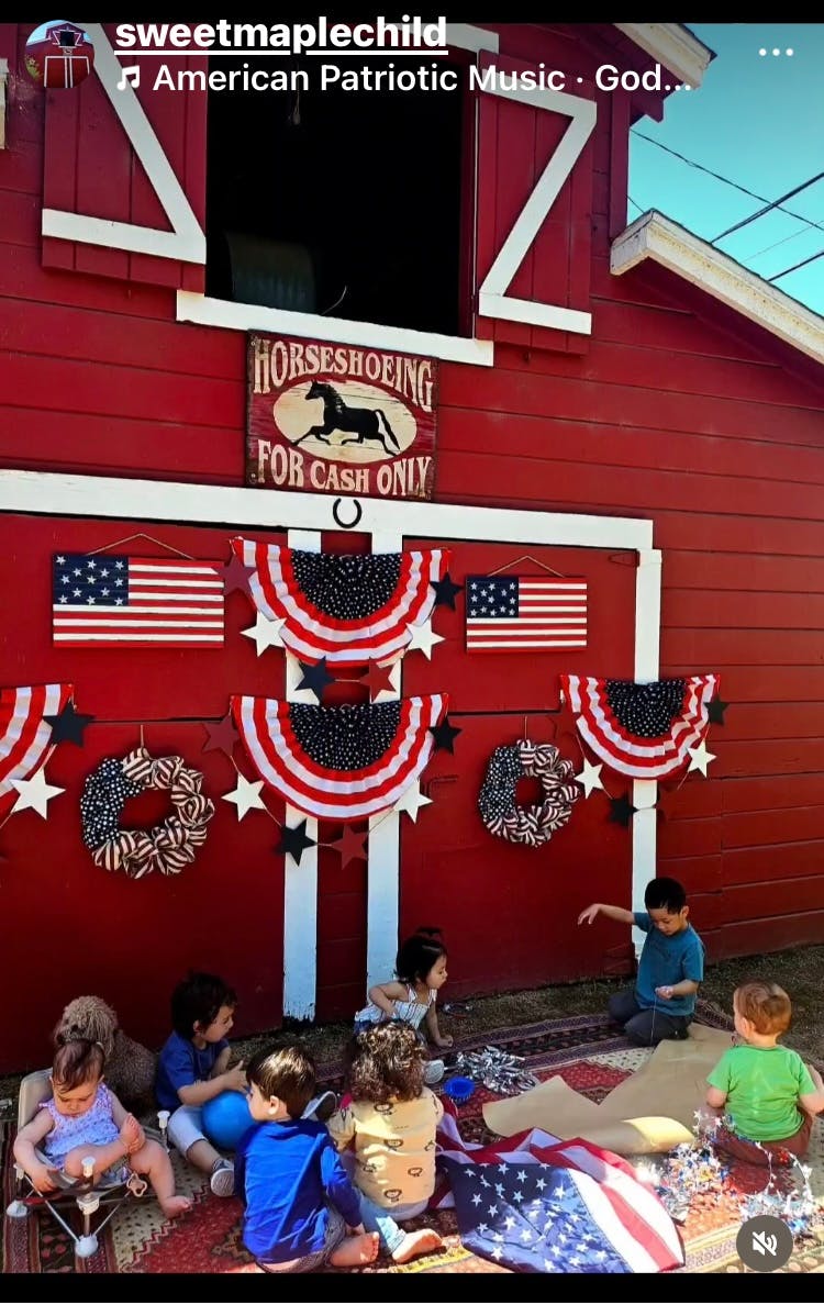 Children sitting on a rug in front of a red barn decorated with patriotic bunting