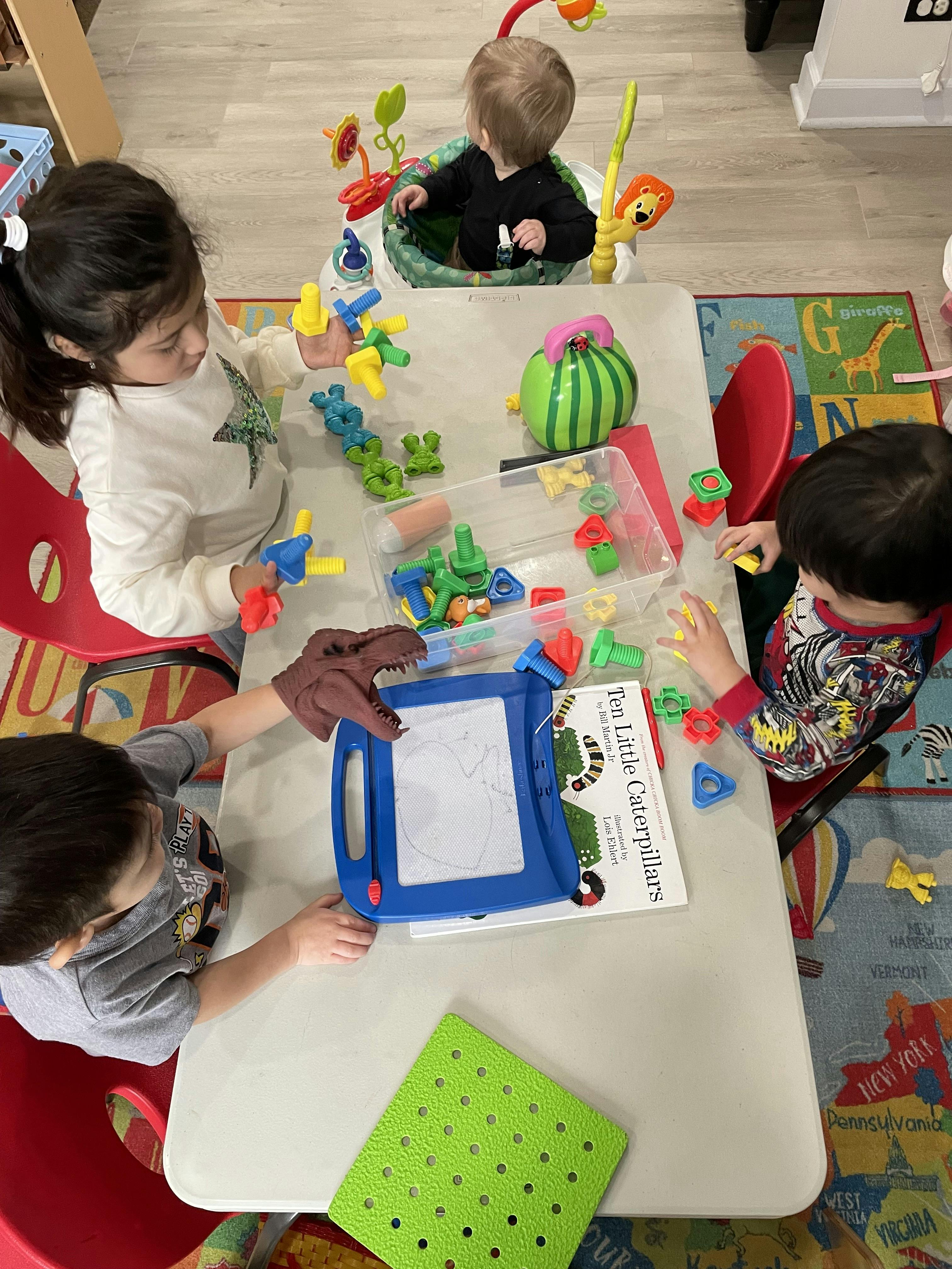 Three children playing with toys at a table, one in a baby bouncer