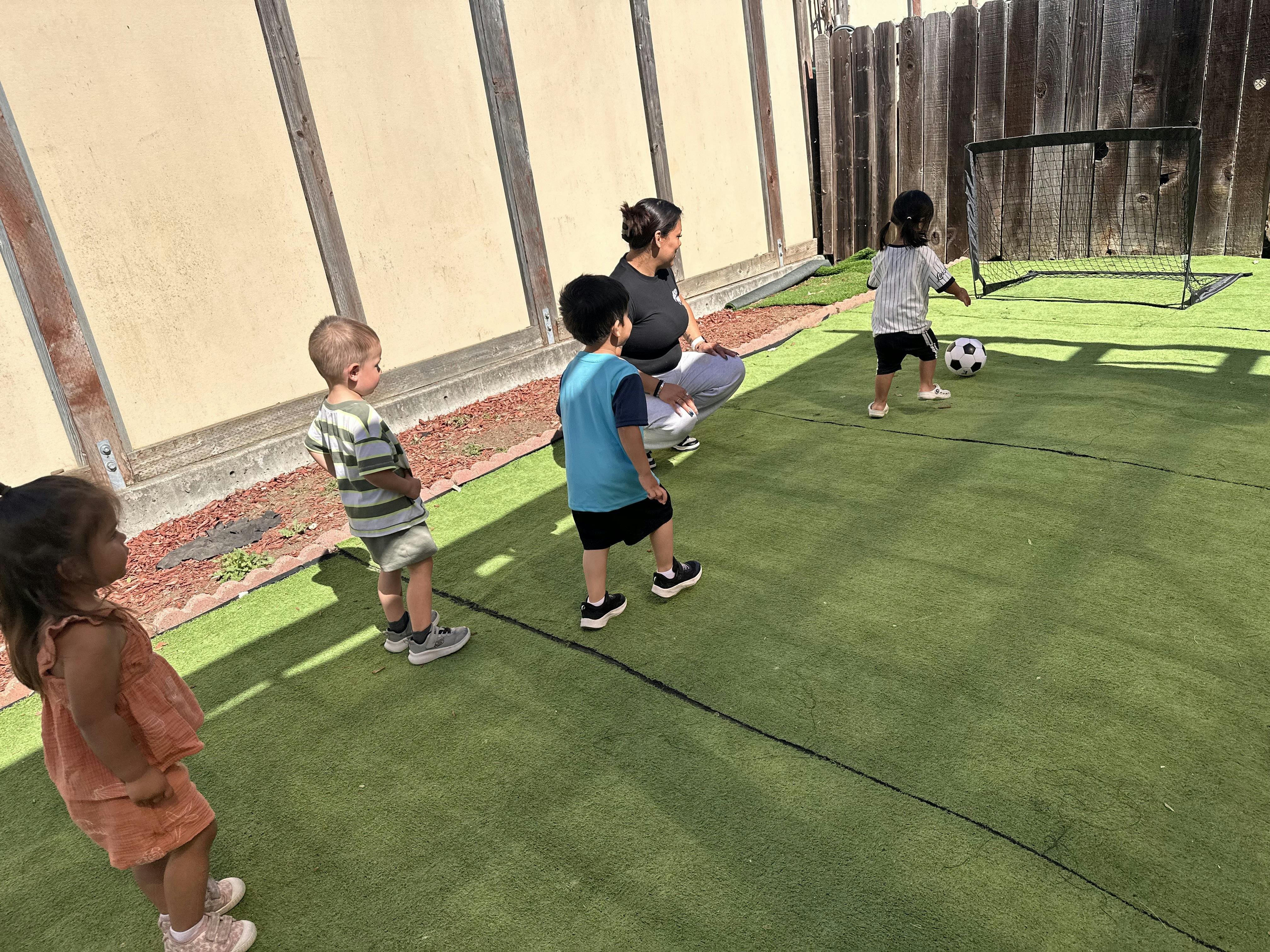 A person observing three children on artificial turf with a soccer goal and ball