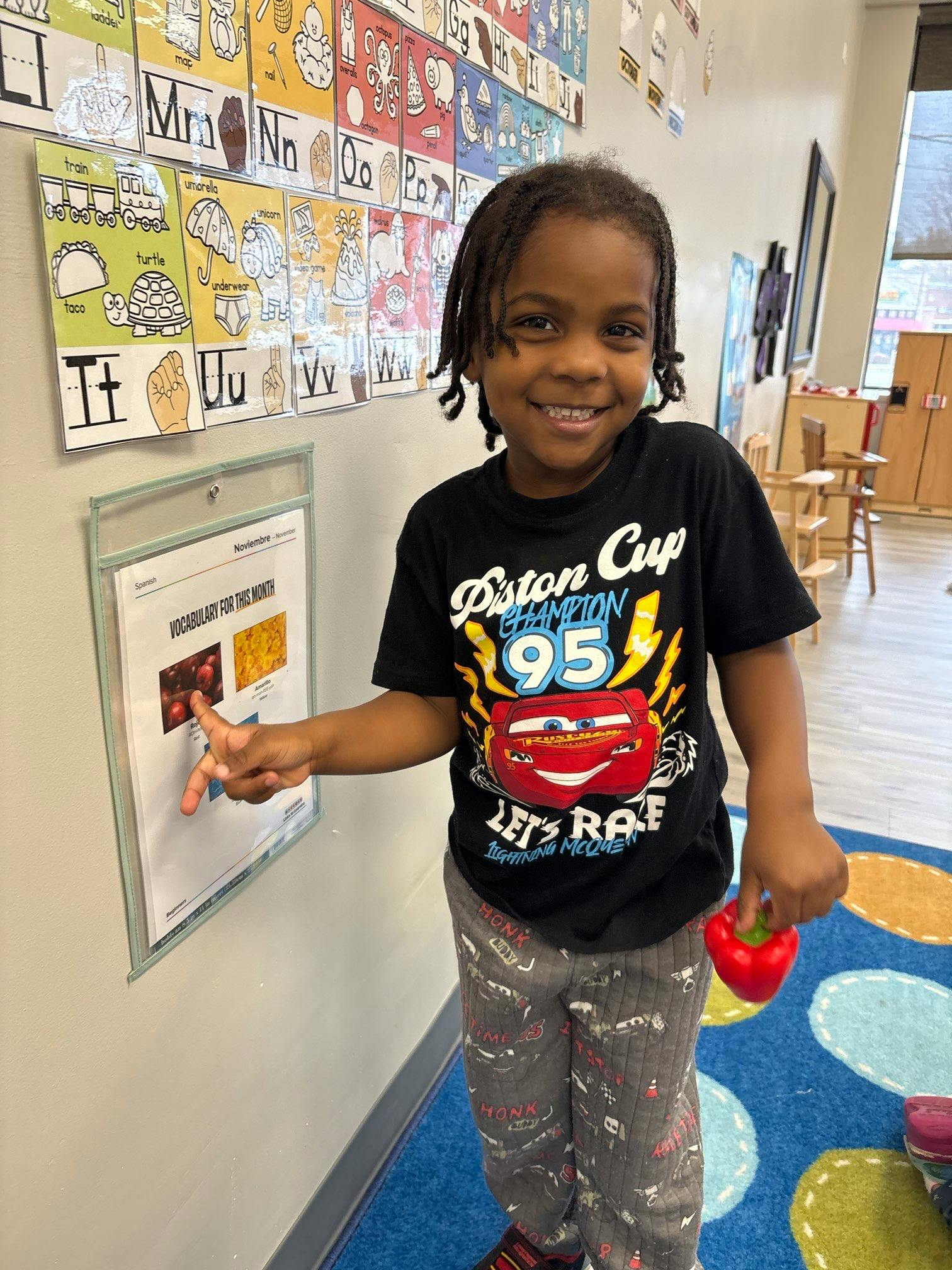 Child pointing at a chart on a wall and holding a red bell pepper