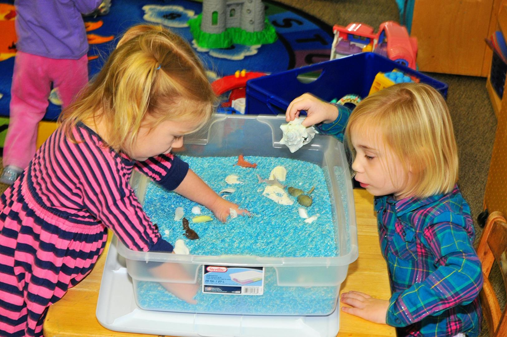 Two children playing with a sensory bin filled with blue beads and small objects
