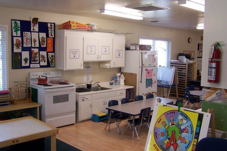 Classroom with a kitchen area, small tables and chairs, and artwork on a bulletin board