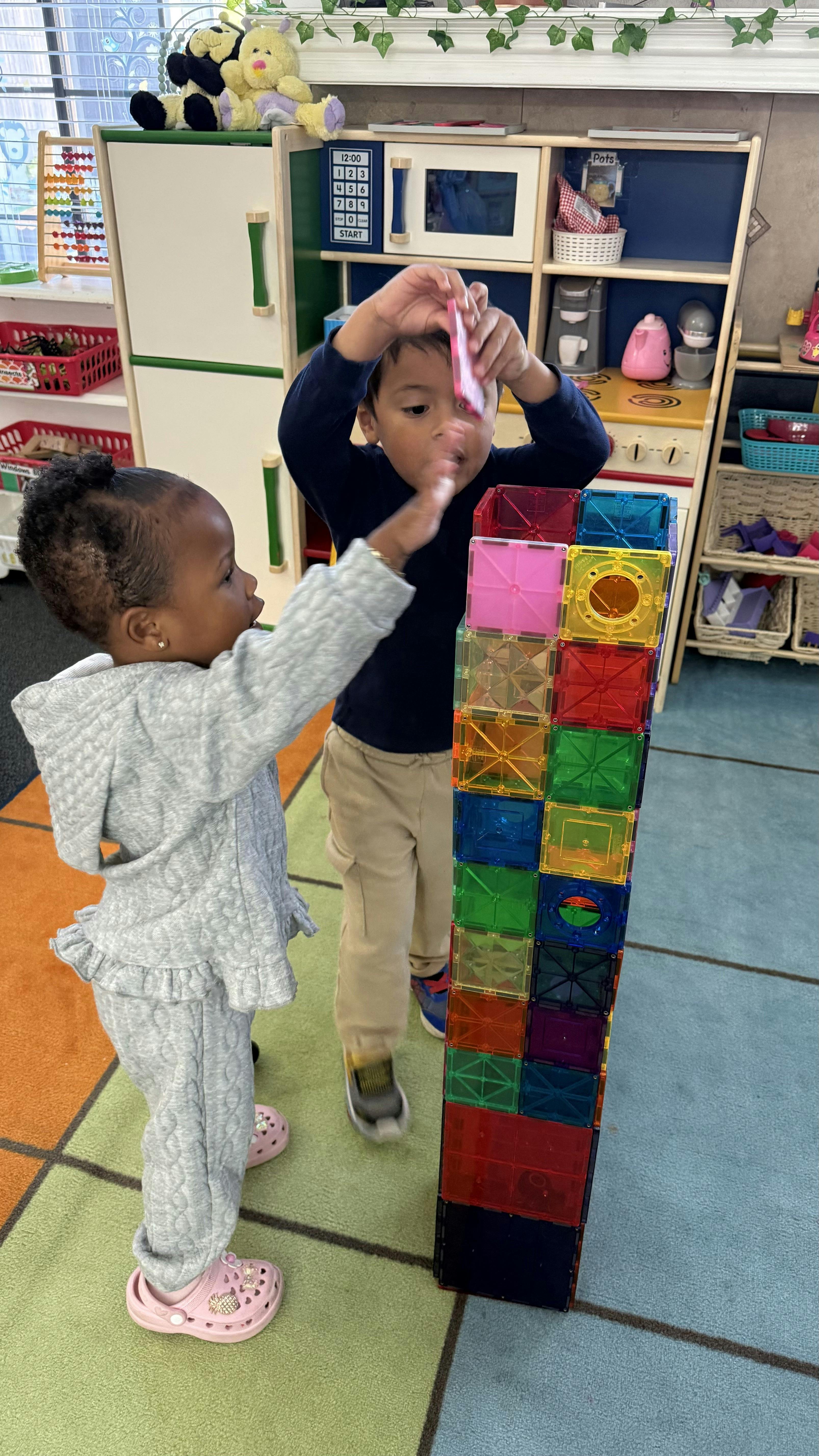 Child adding magnetic tile to a tall tower while another child watches