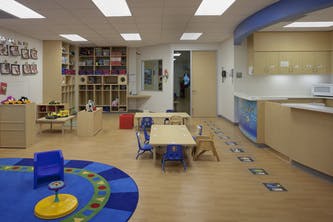 Empty classroom with small tables and chairs, play area with blue rug