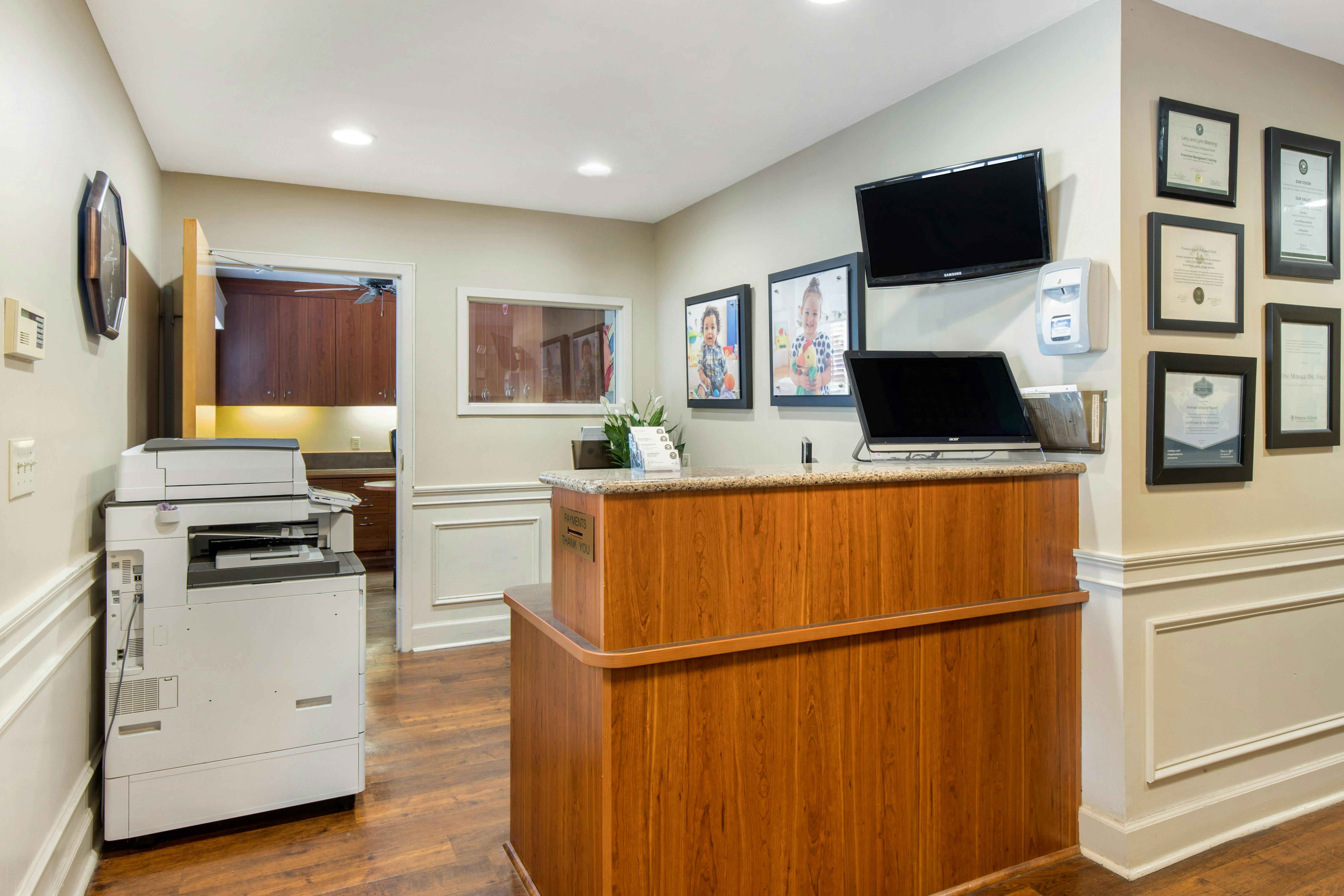 Reception area with a wooden desk, monitors, framed photos of children, and diplomas