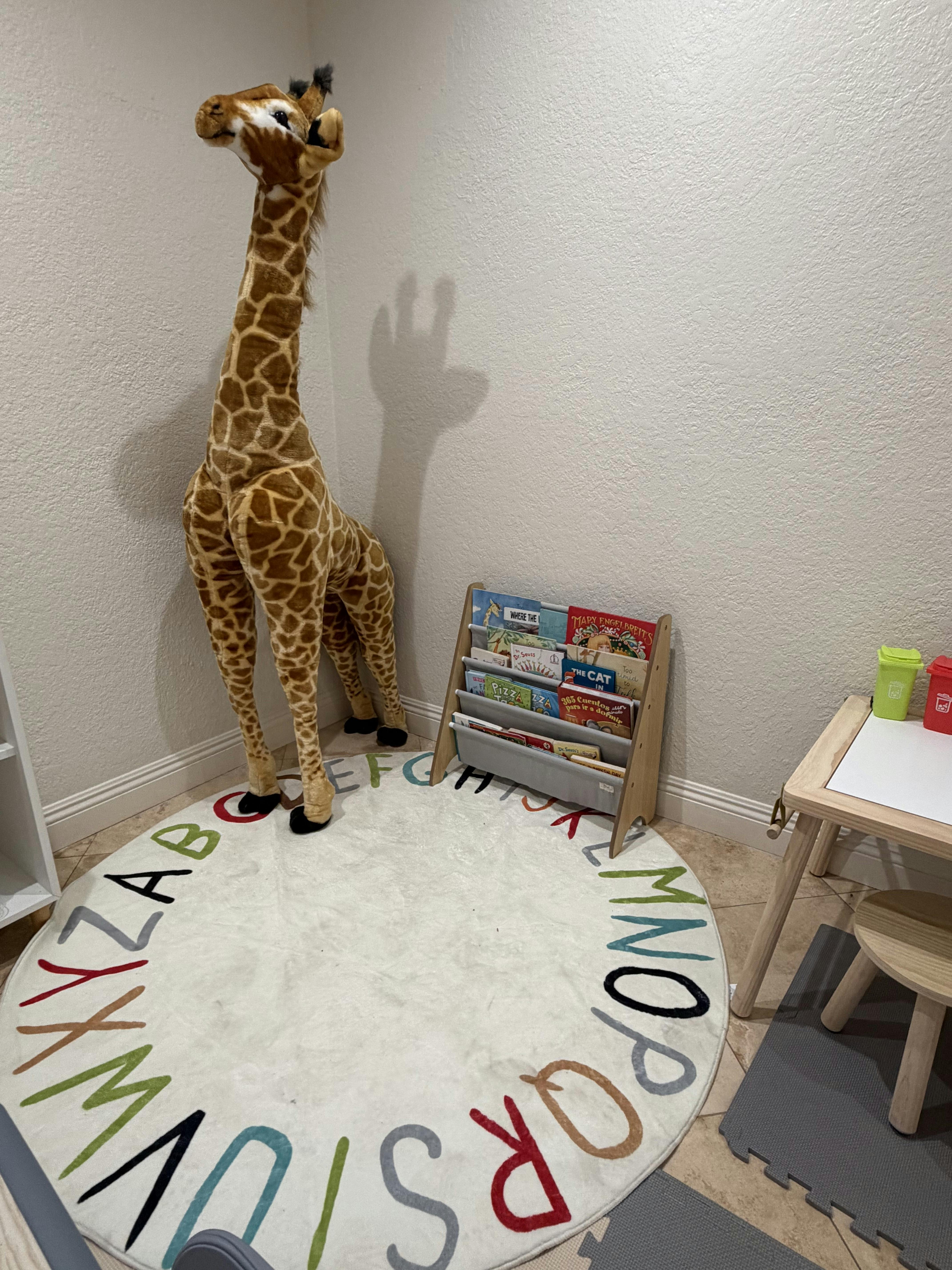 Large stuffed giraffe standing on an alphabet rug near a book rack