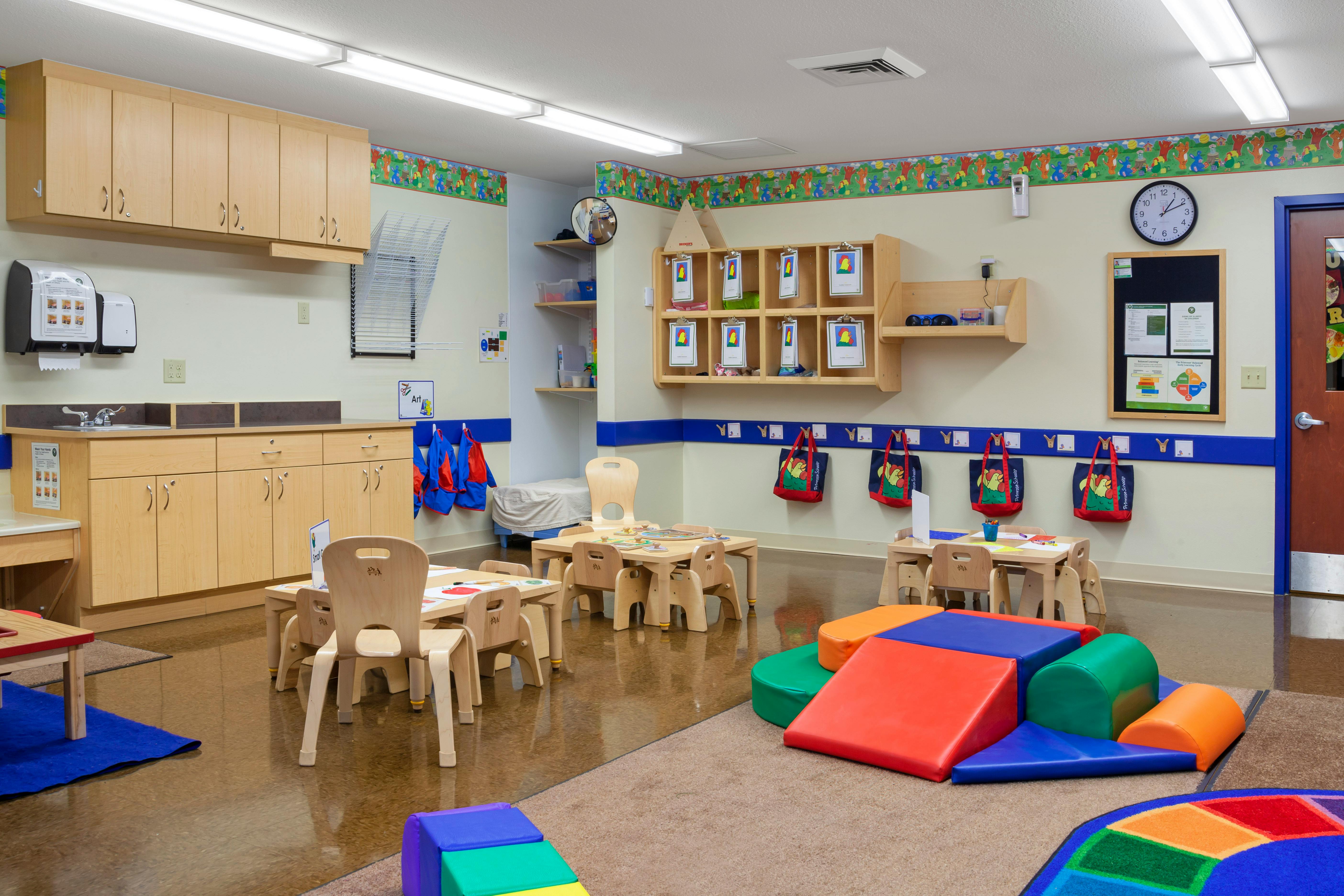 An empty classroom with small tables and chairs, a sink counter, and play mats