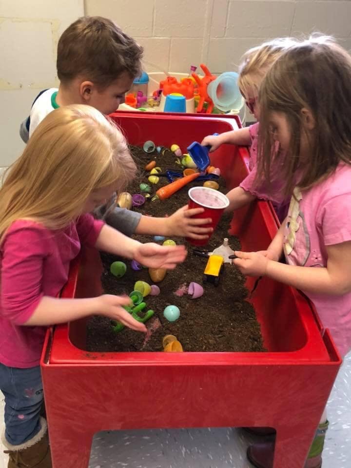 Children playing in a sensory bin with dirt and small toys