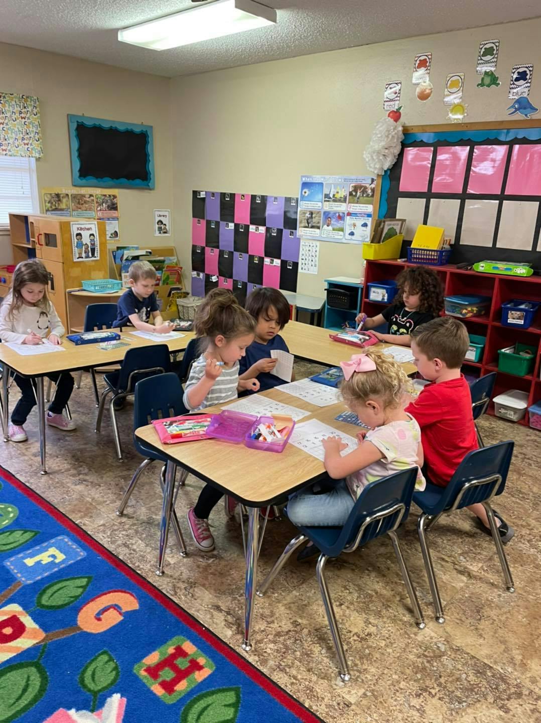 Children sitting at tables in a classroom, writing and drawing