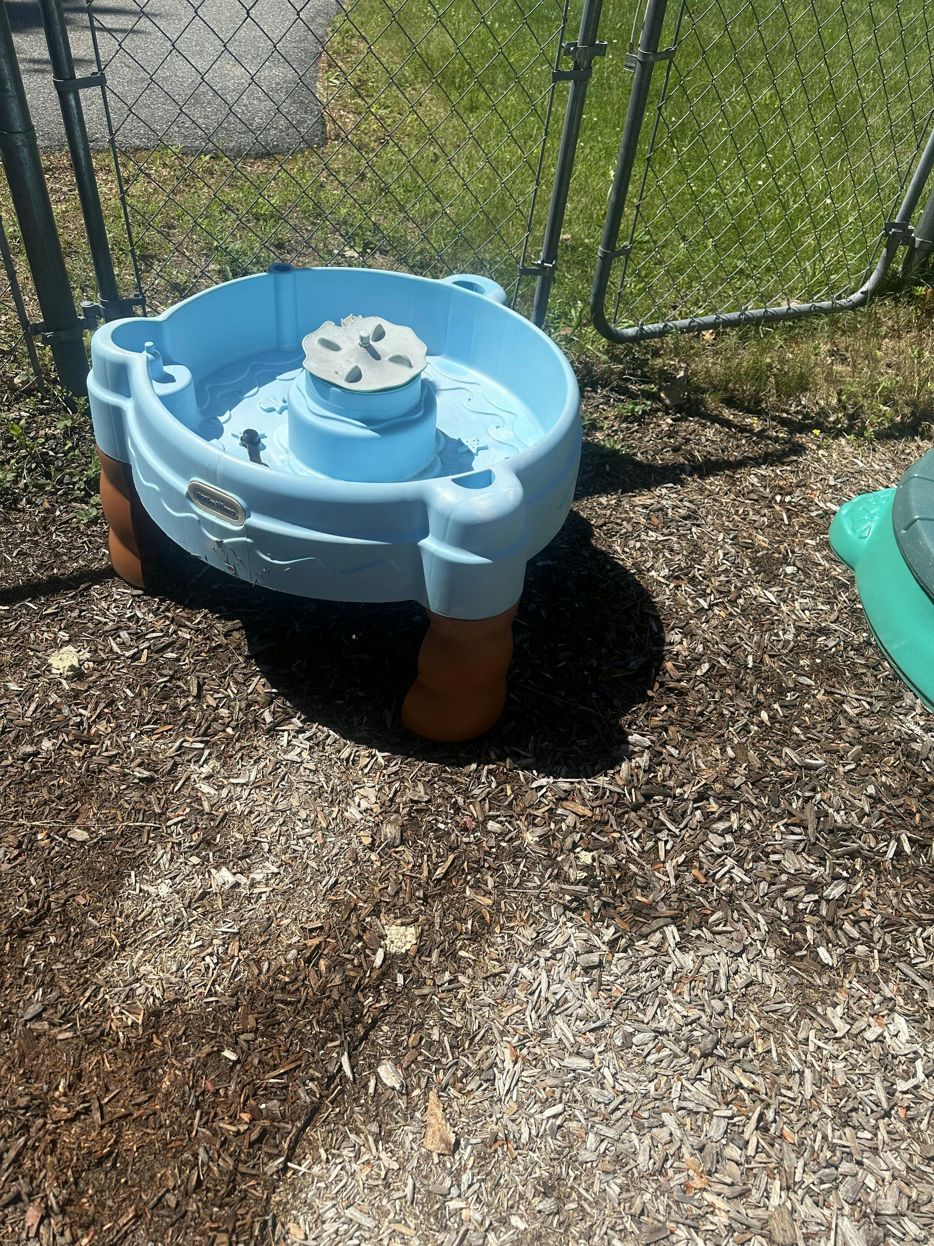 Plastic water play table on a wood chip surface near a chain link fence
