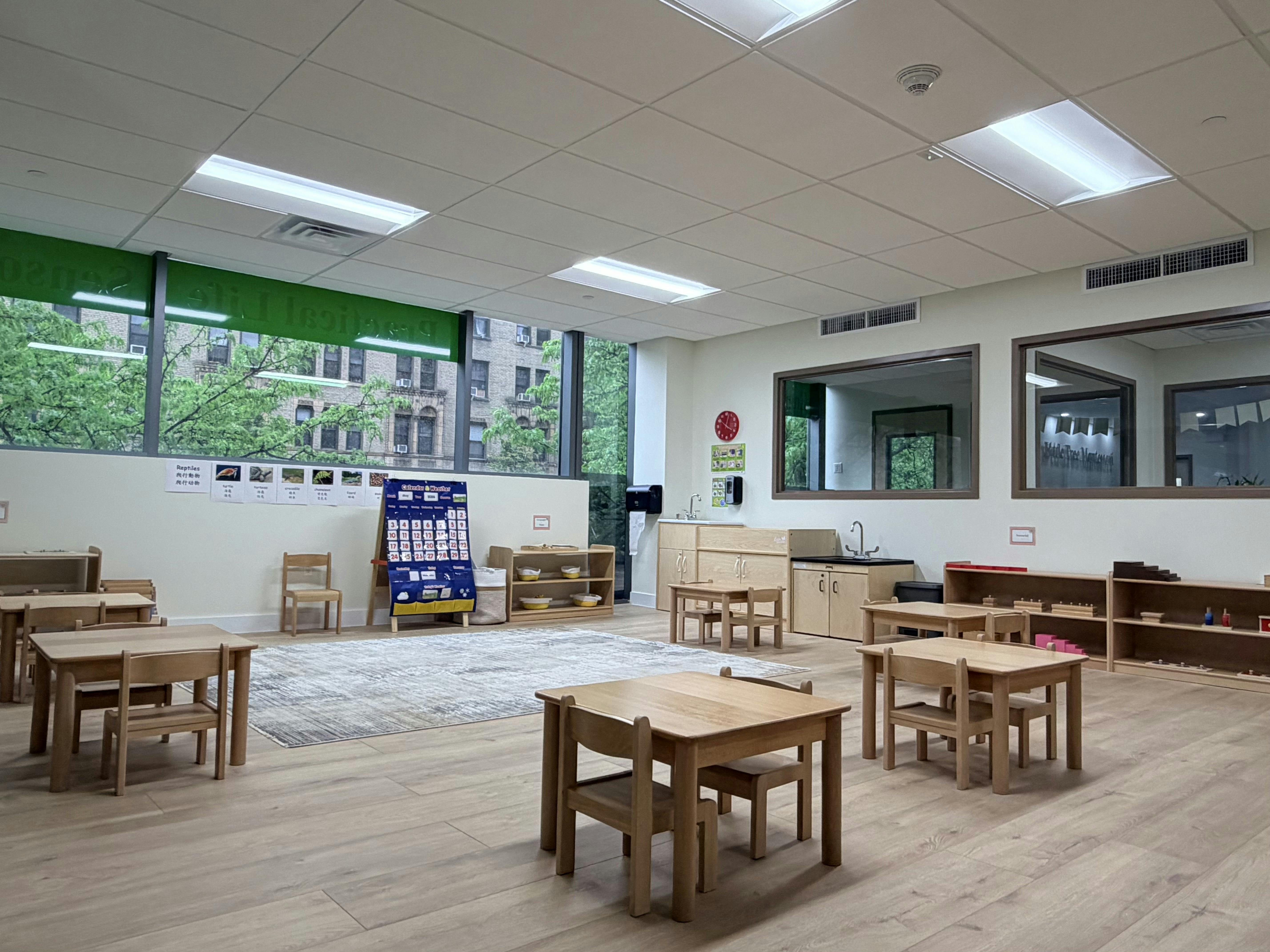 Empty classroom with small wooden tables and chairs, large windows, and learning materials