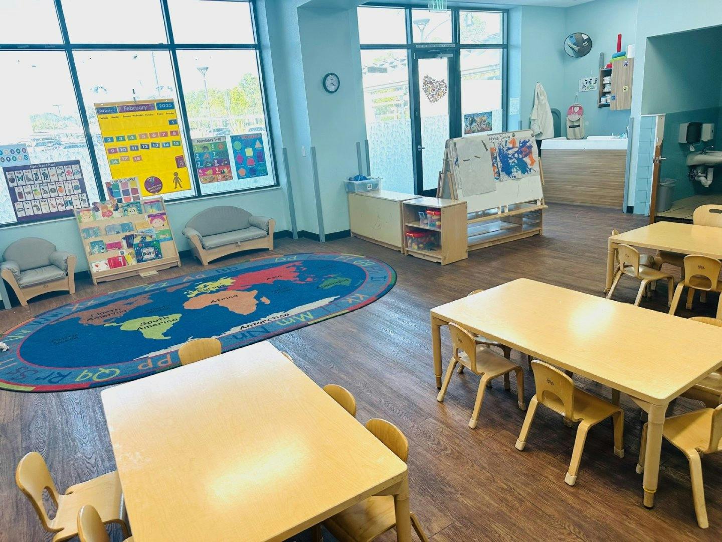 A classroom with small tables and chairs, a world map rug, and windows