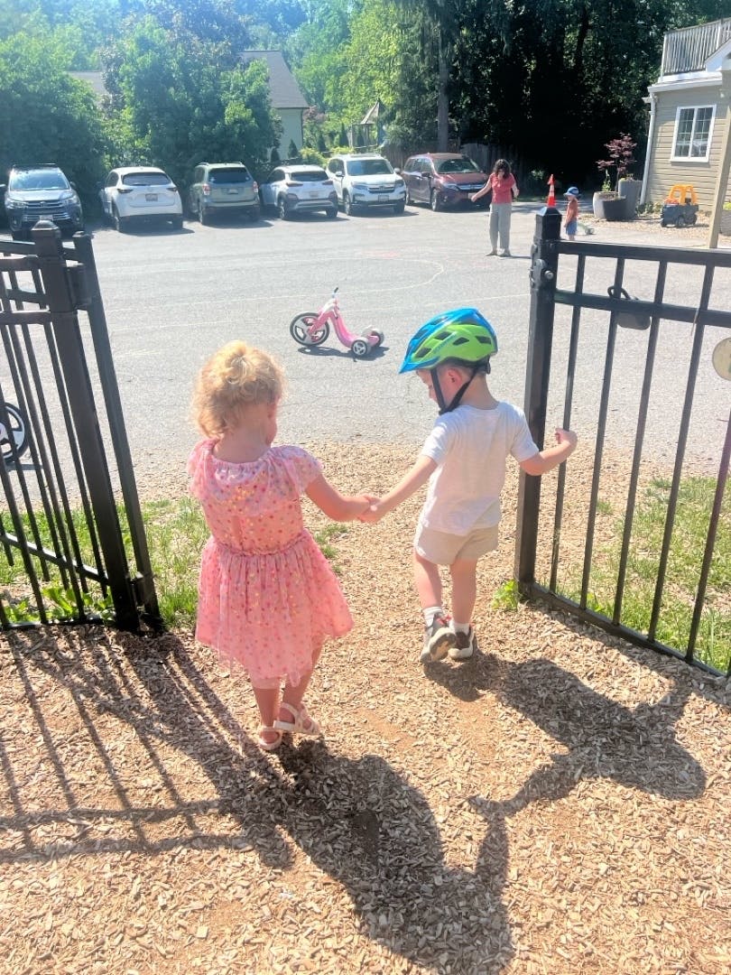 Two children holding hands walking through a gate toward a parking lot