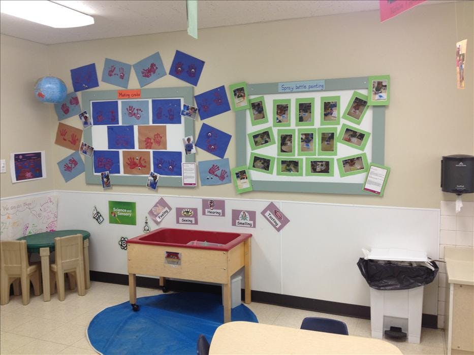 Classroom wall with child handprint art, photos of children painting, and a sensory table