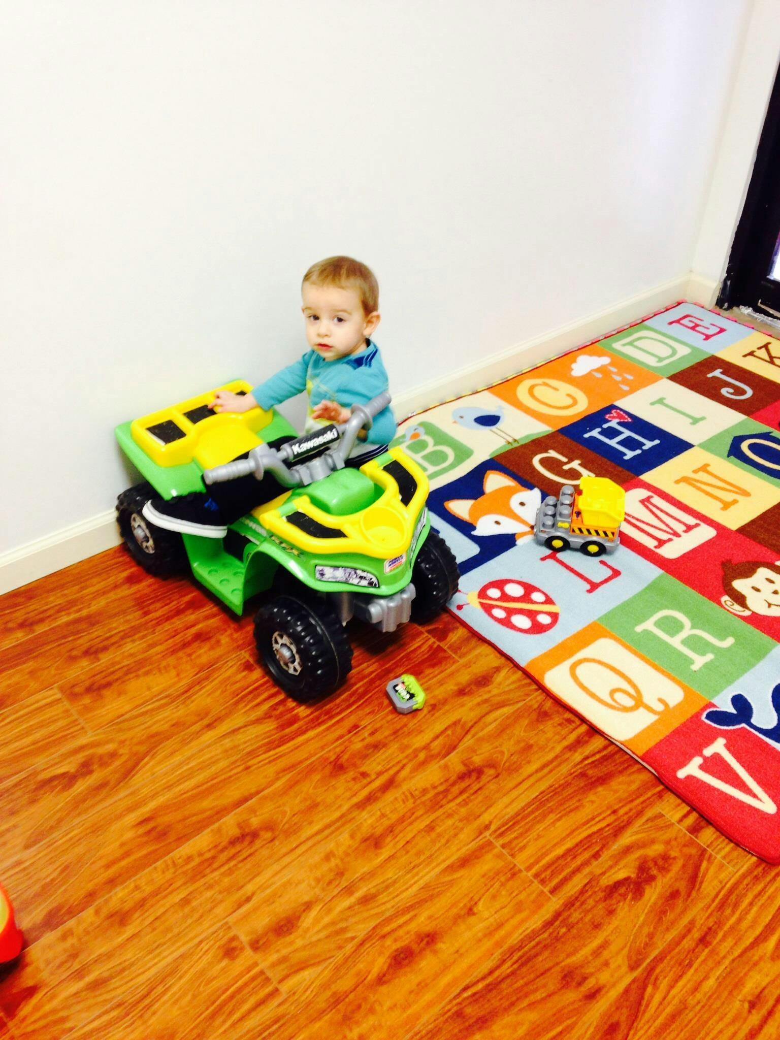 Child on a toy ATV on a wooden floor next to an alphabet rug