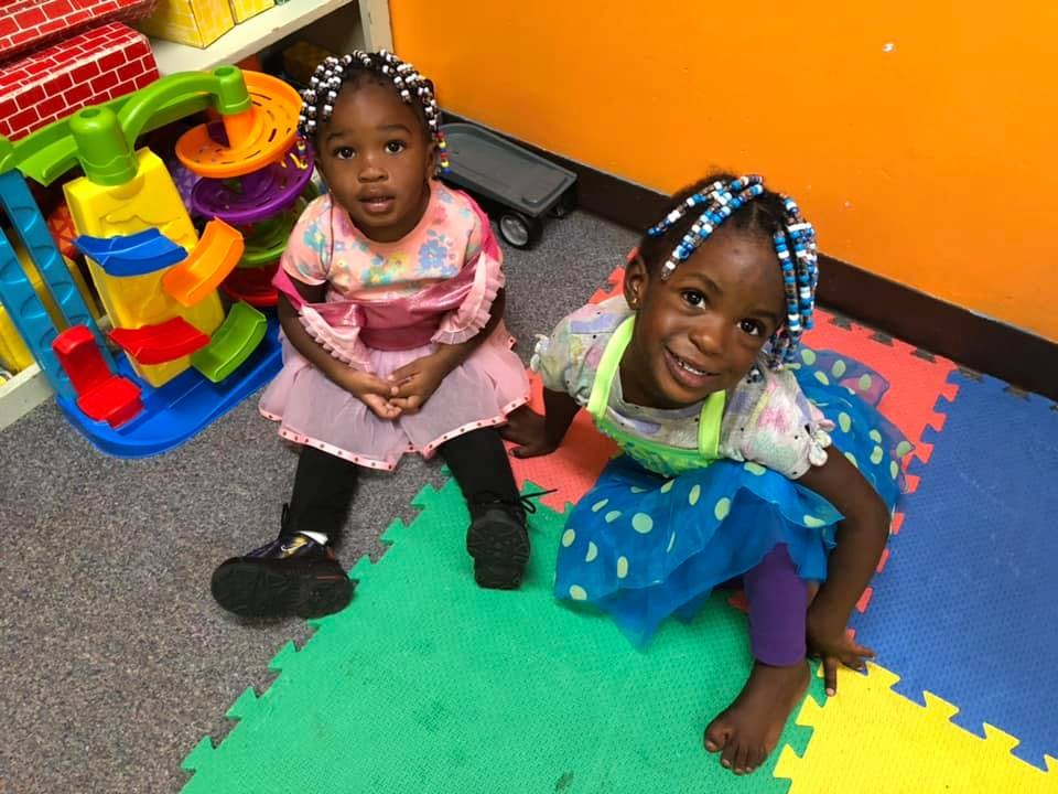 Two children sitting on a colorful mat in a room with toys