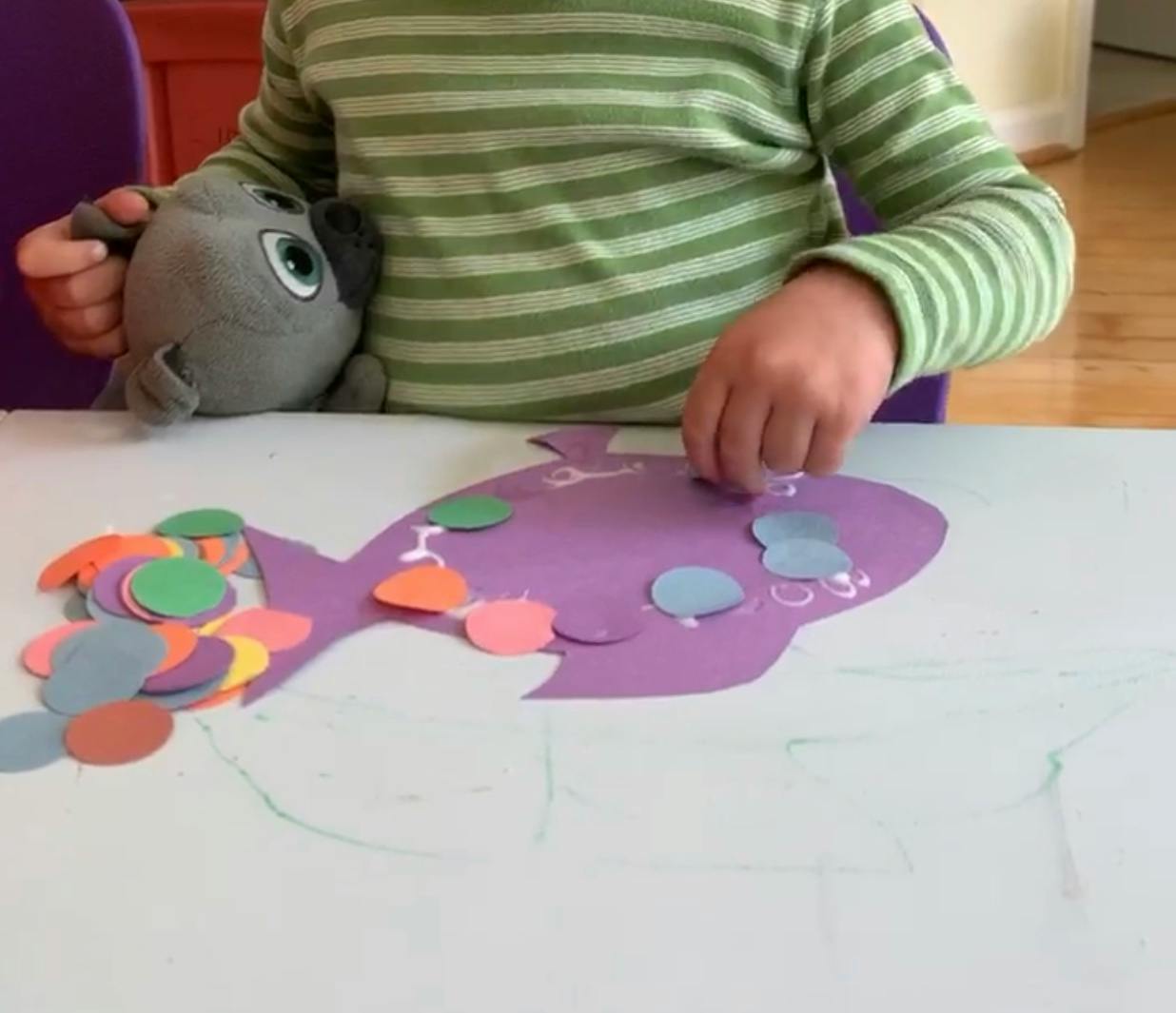 Child making a craft with a purple fish cutout and colorful paper circles