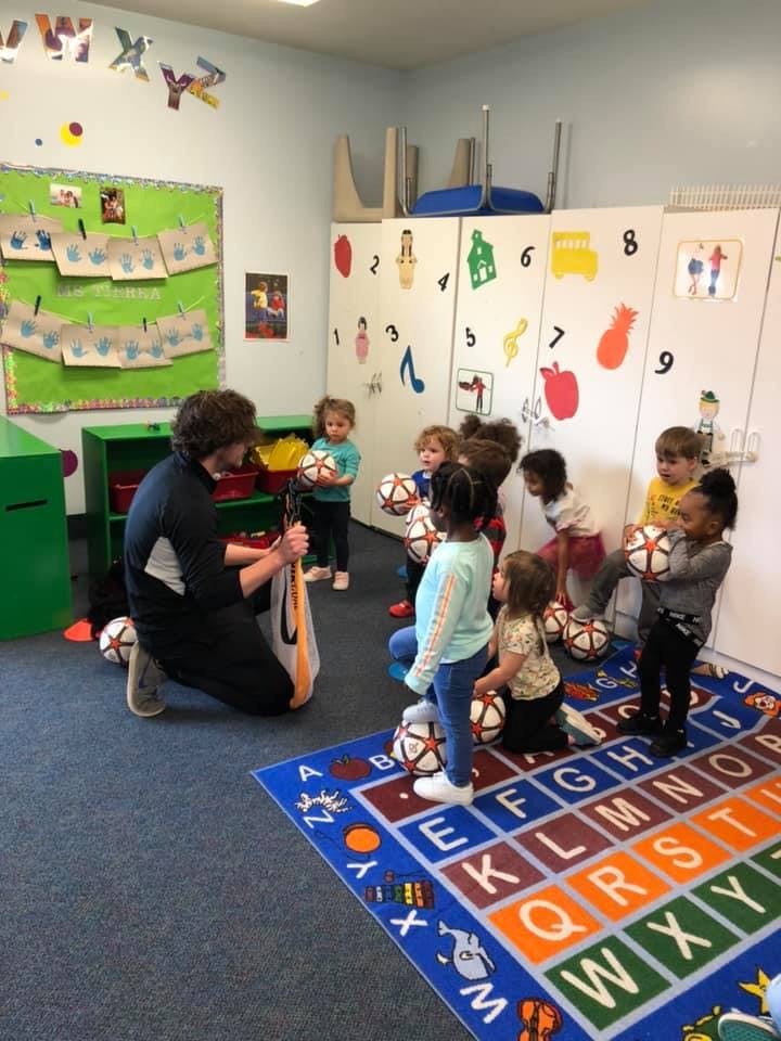 Adult kneeling with children holding soccer balls in a classroom