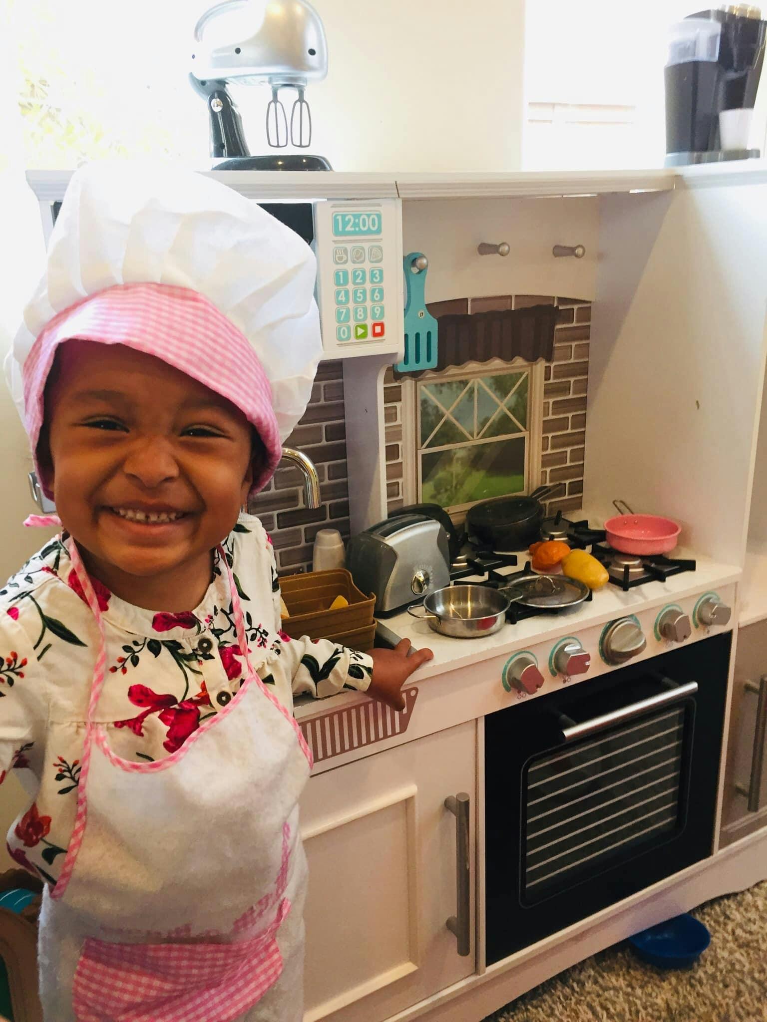 Child in chef hat and apron standing in front of a play kitchen