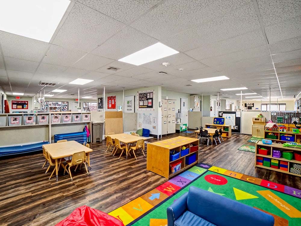 Empty classroom with small tables and chairs, storage, and colorful play rug