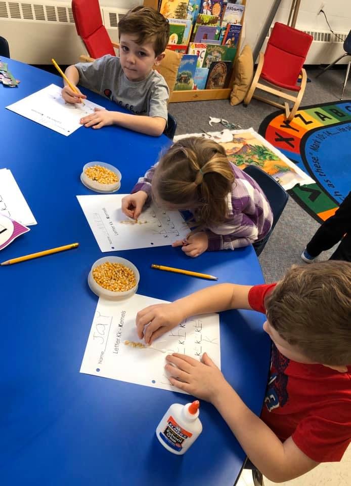 Three children working on crafts at a blue table in a classroom