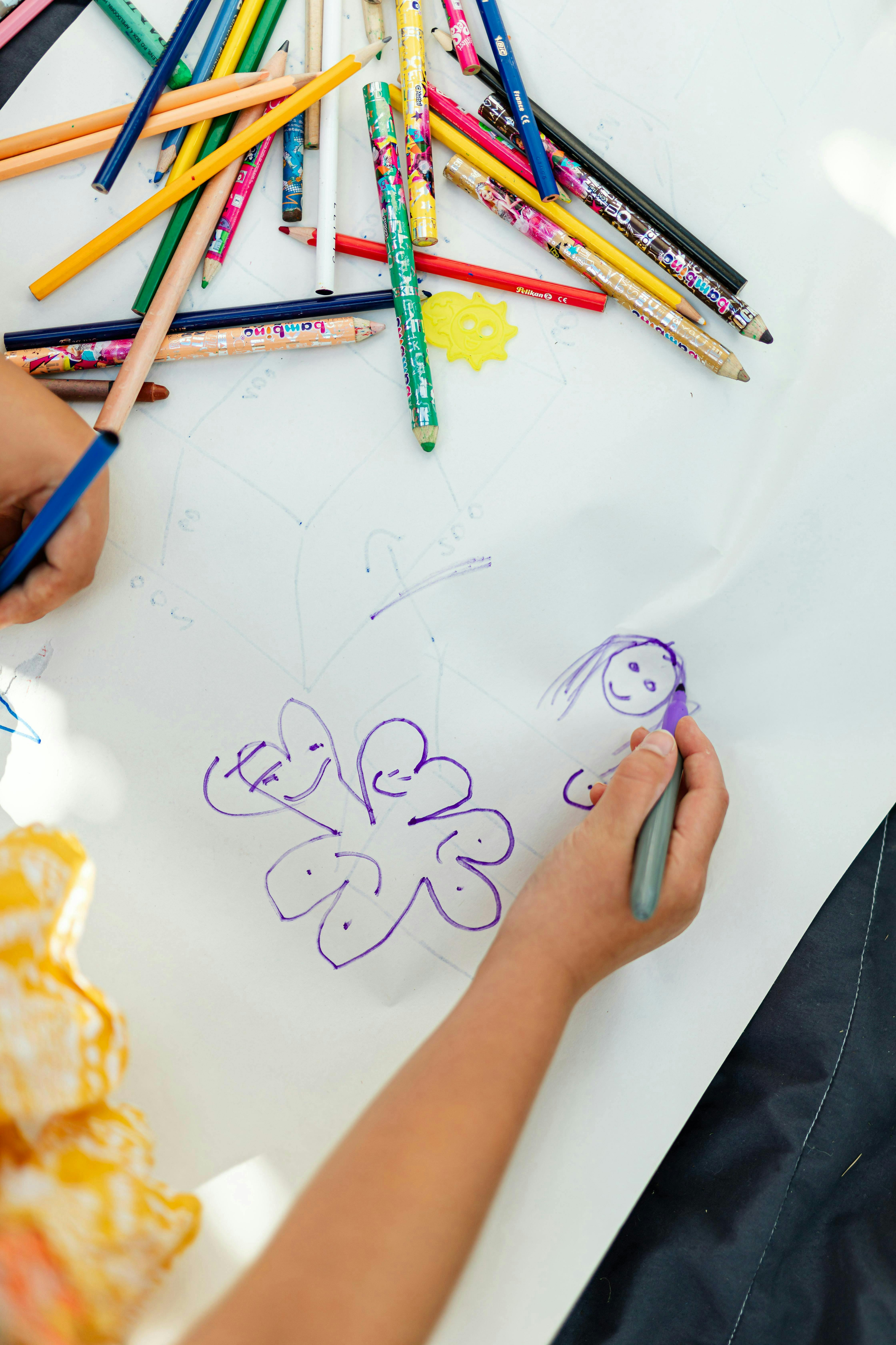 Child's hands drawing on white paper with a purple marker, colored pencils scattered