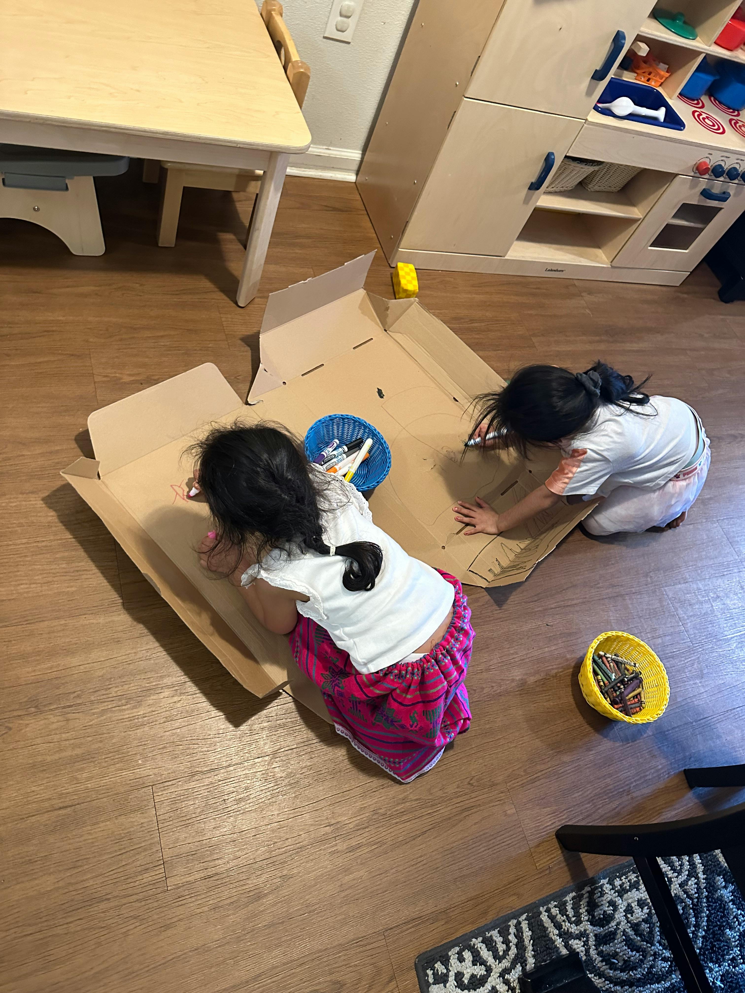 Two children drawing on a cardboard box on the floor