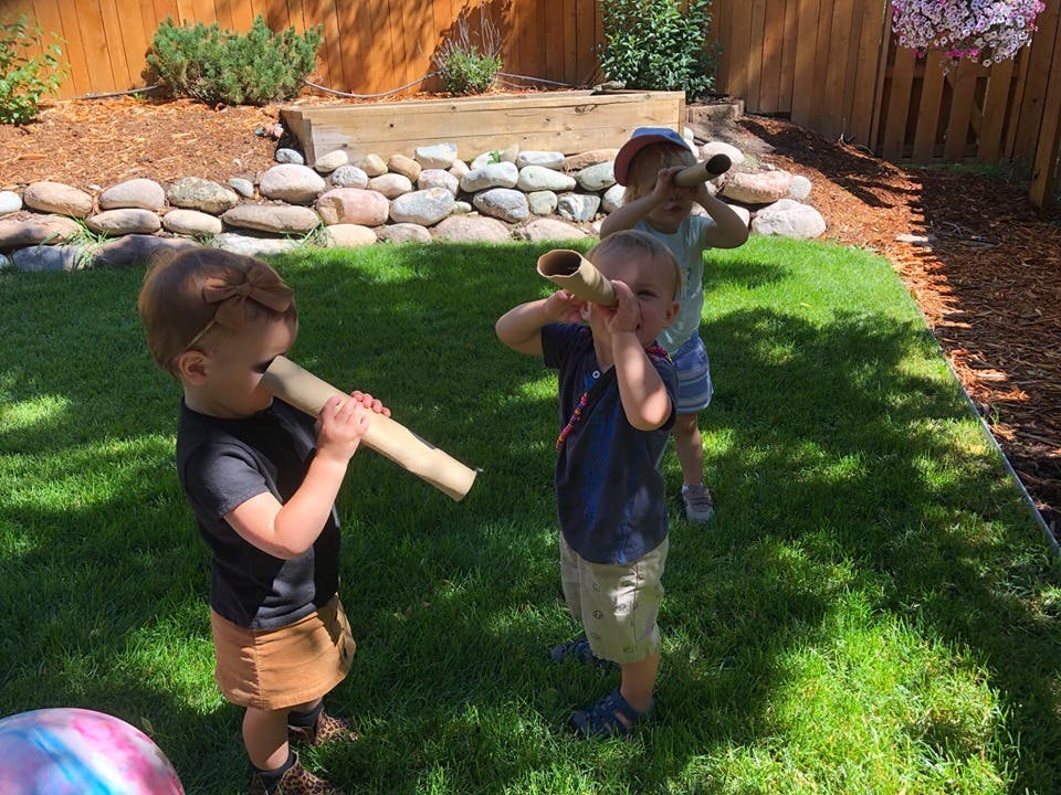 Three children playing with cardboard tubes in a grassy yard