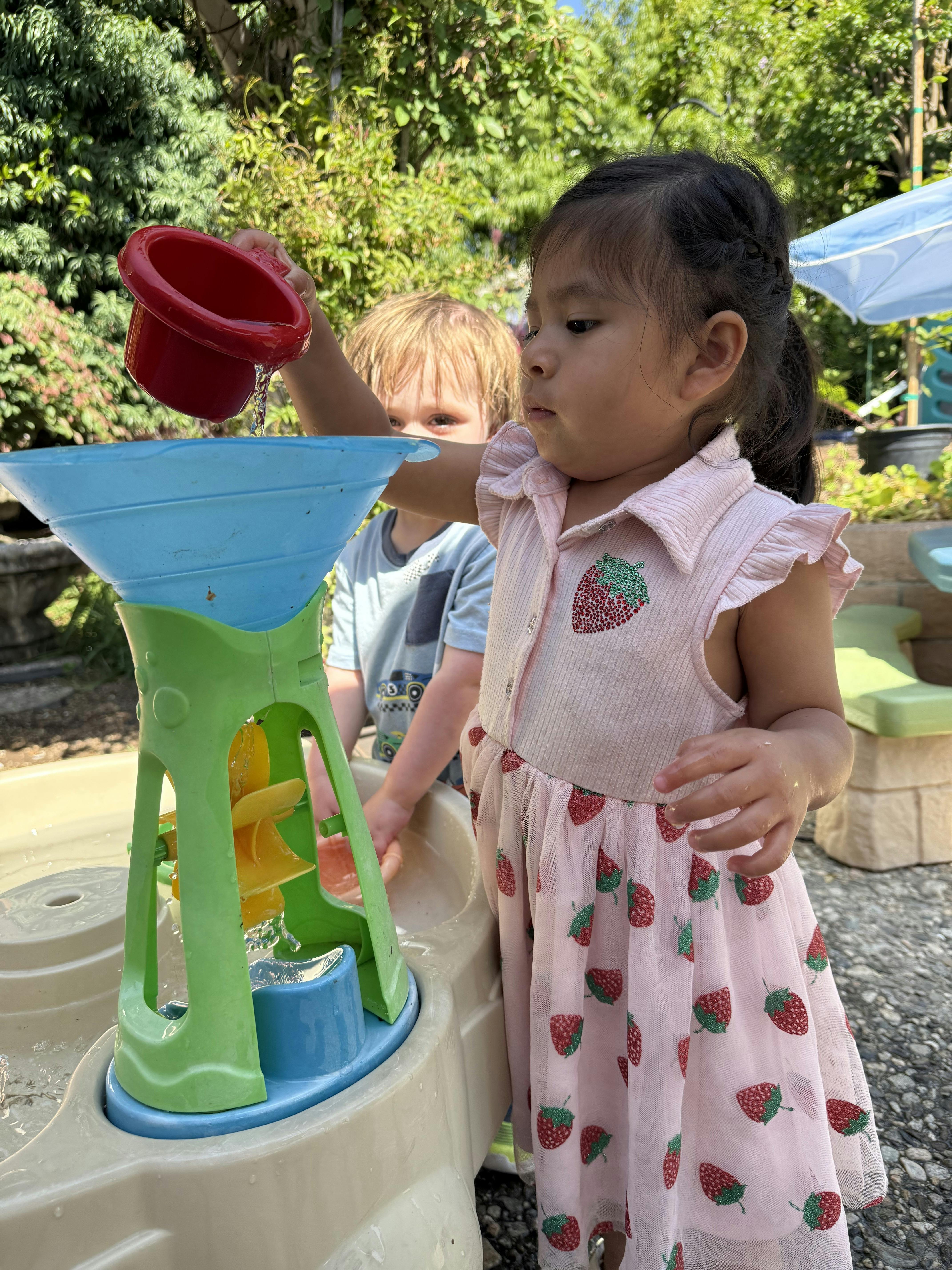 Child pouring water into a water table toy with another child in the background