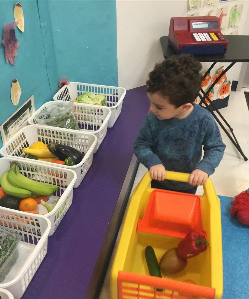 Child pushing a toy shopping cart past baskets of produce