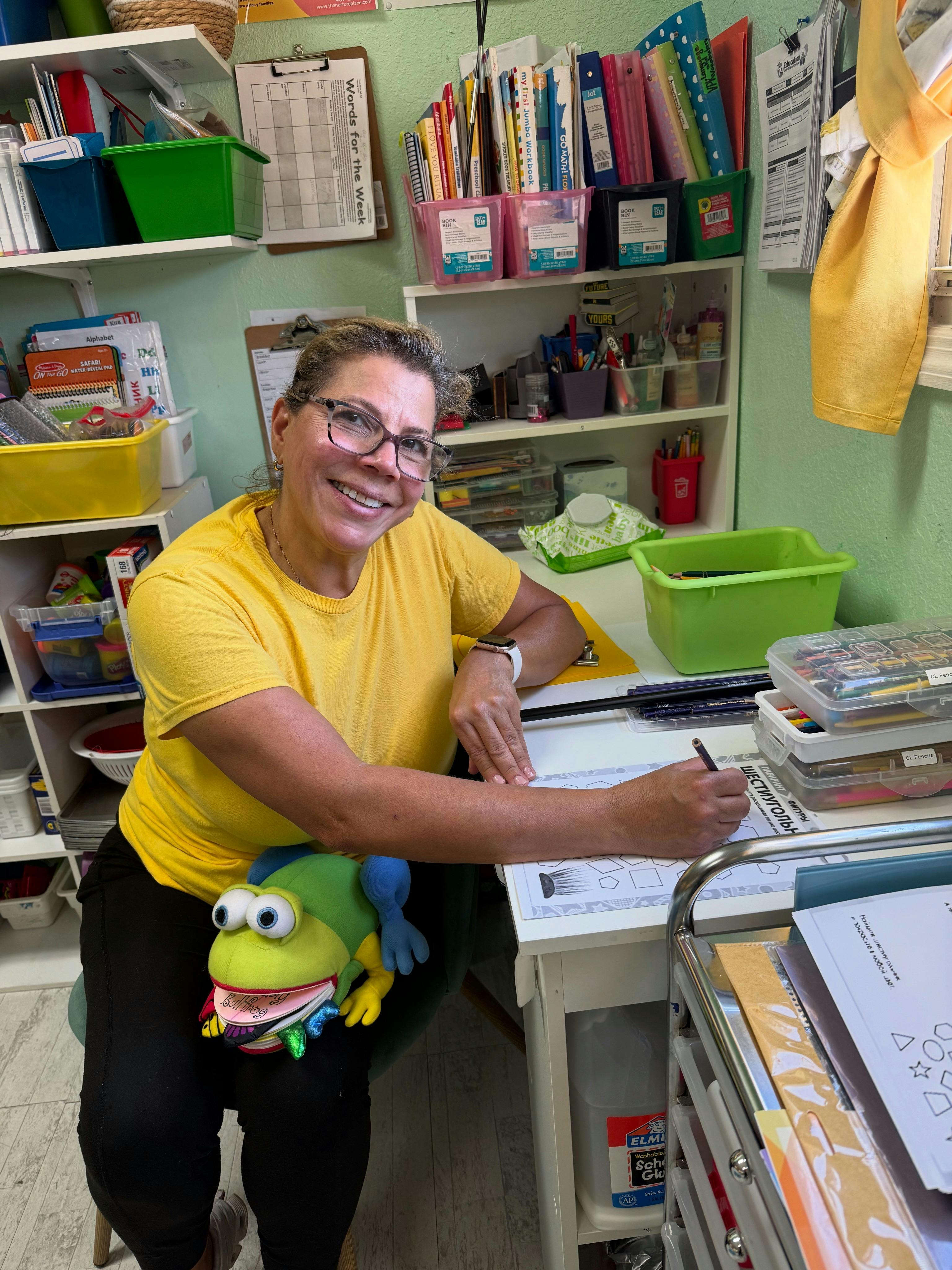 Person in glasses writing at a desk with a frog puppet on their lap