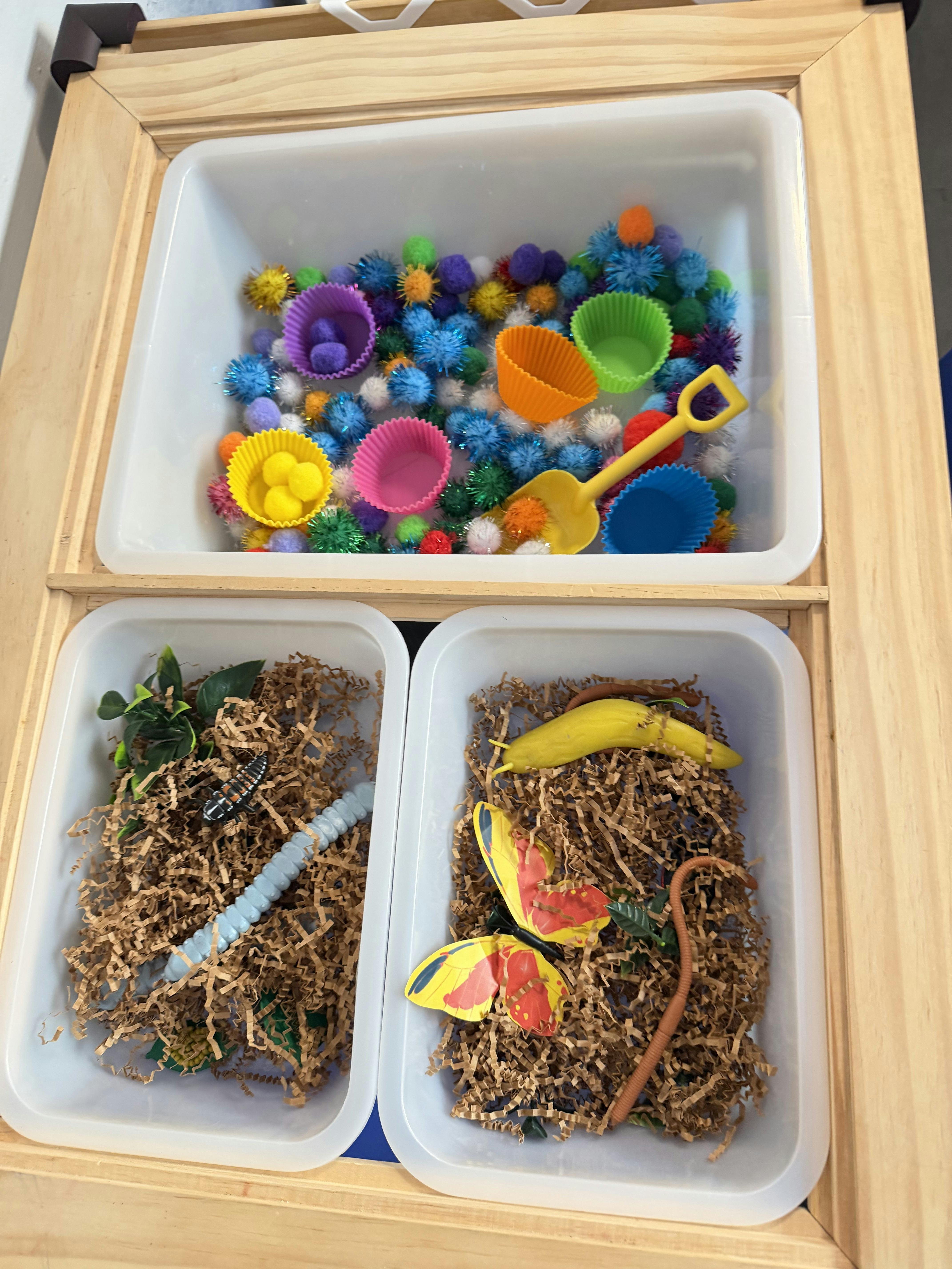 Sensory bin with pom-poms and cups, and two bins with shredded paper and bug toys