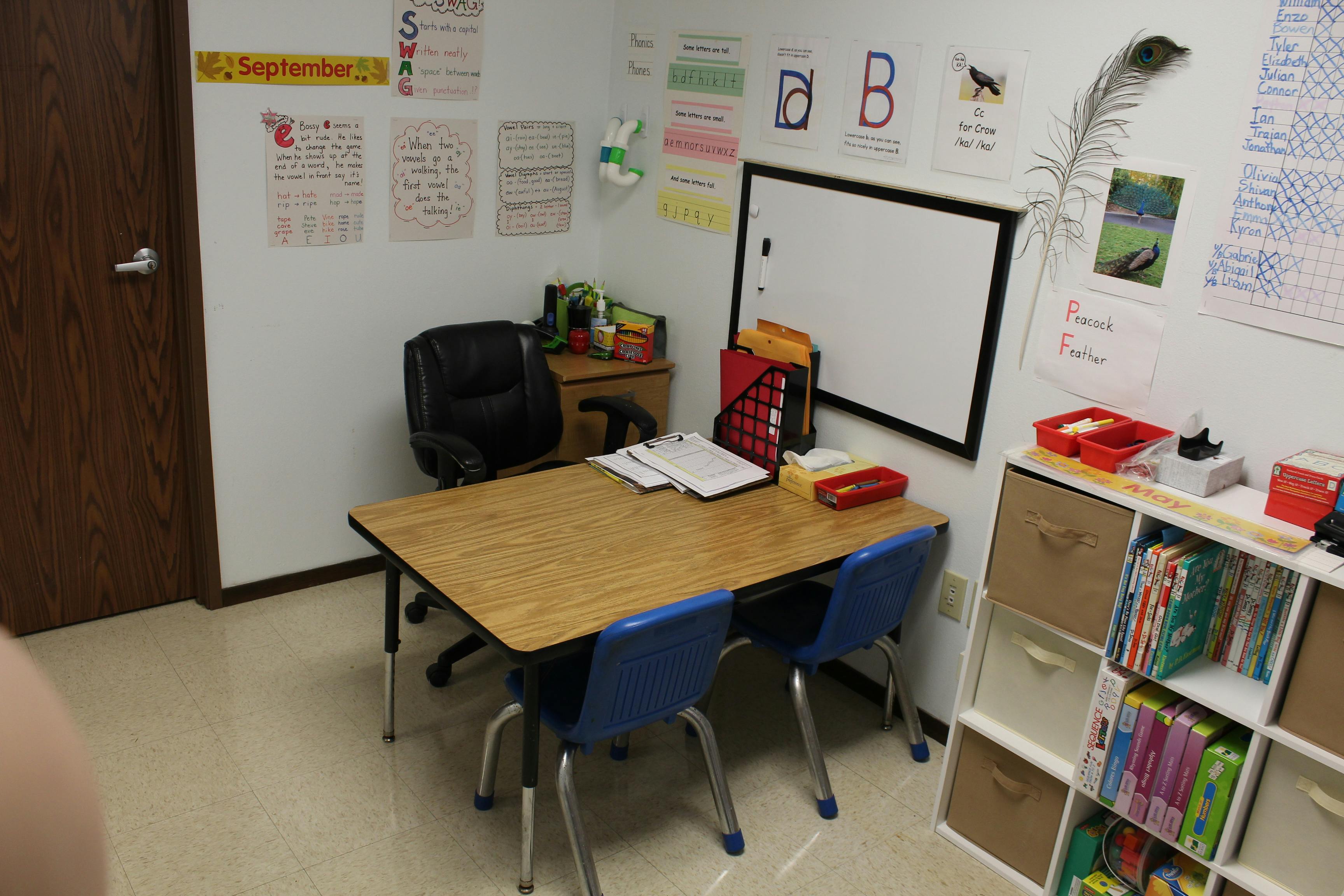 Classroom with a wooden table, blue chairs, black office chair, and educational posters on the wall