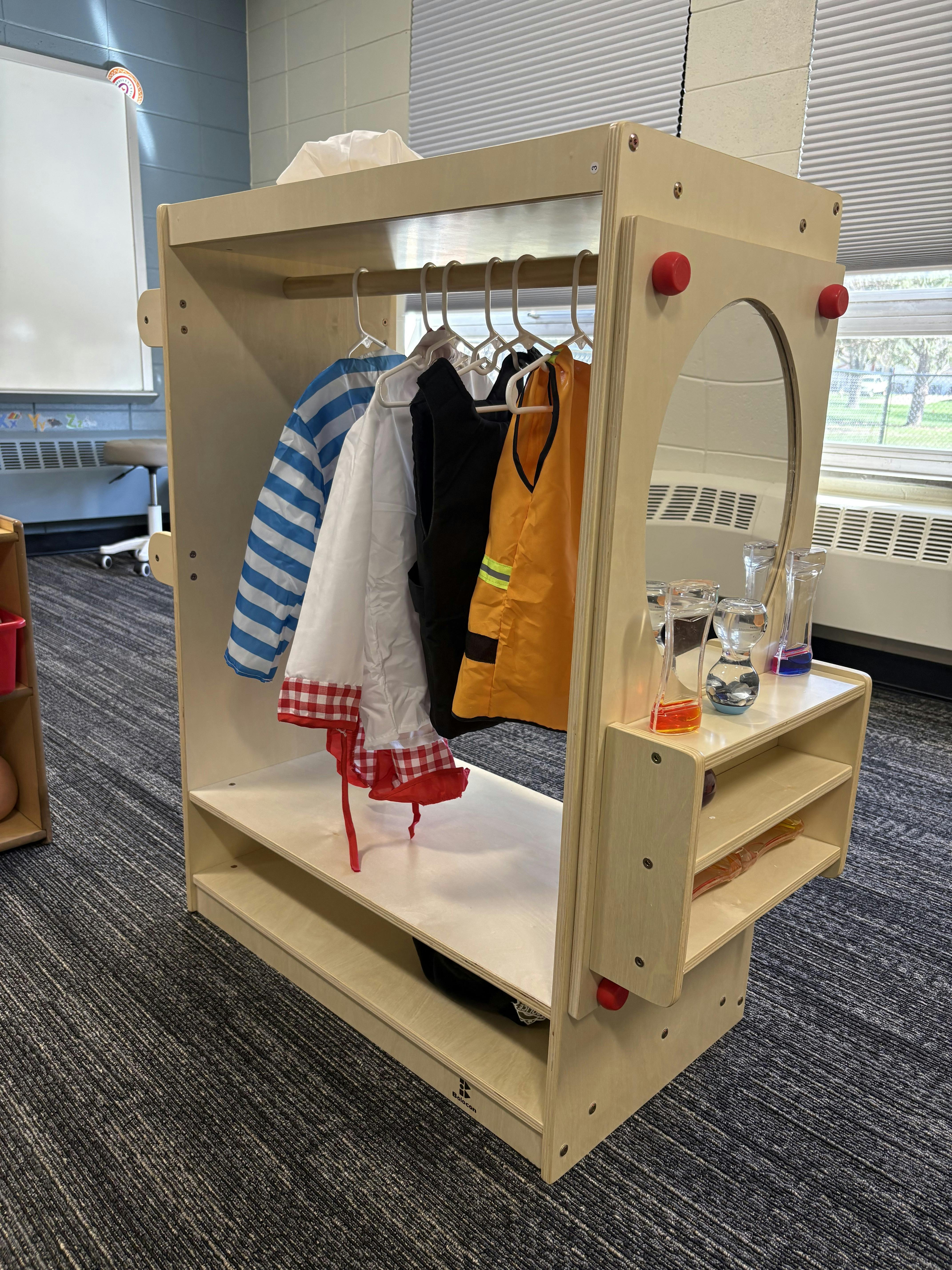 Dress-up clothes on a wooden rack with a mirror and sensory bottles in a classroom