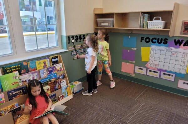 Three children in a classroom, two standing and one reading next to a bookshelf