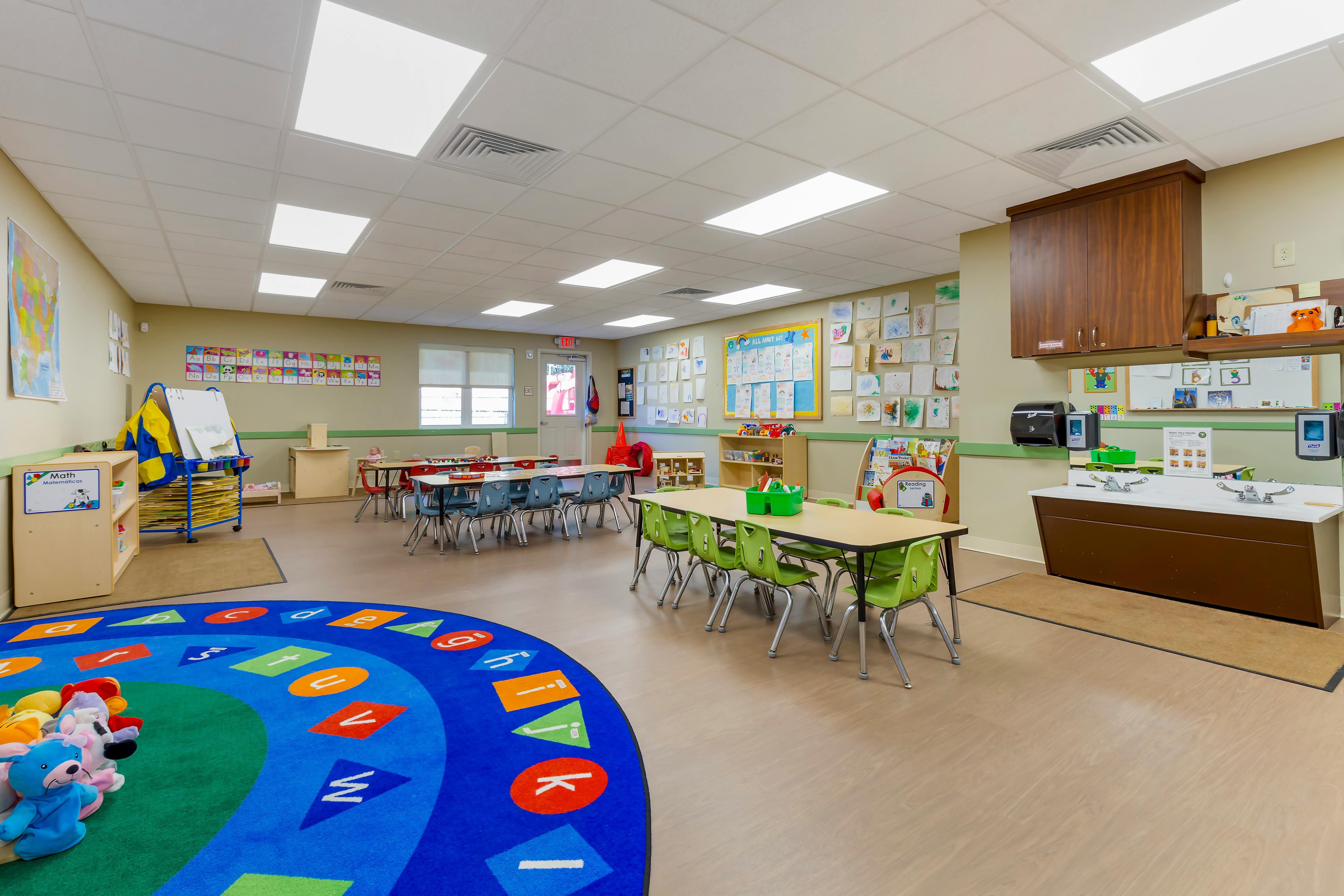 Classroom with small tables and chairs, a circular alphabet rug, and a handwashing station