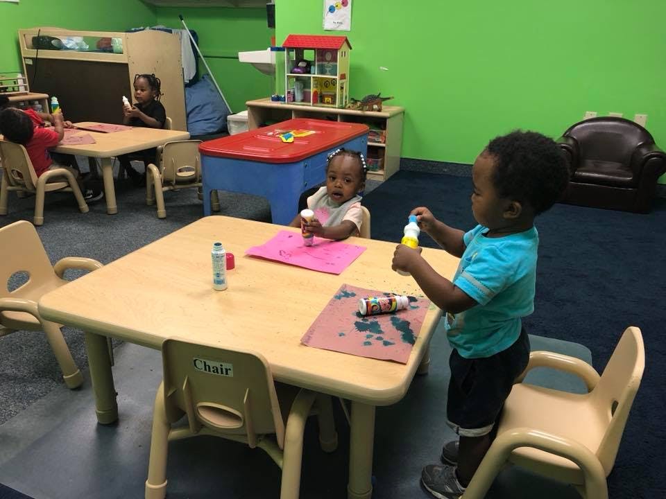 Children at small tables in a classroom, one child standing and holding a bottle