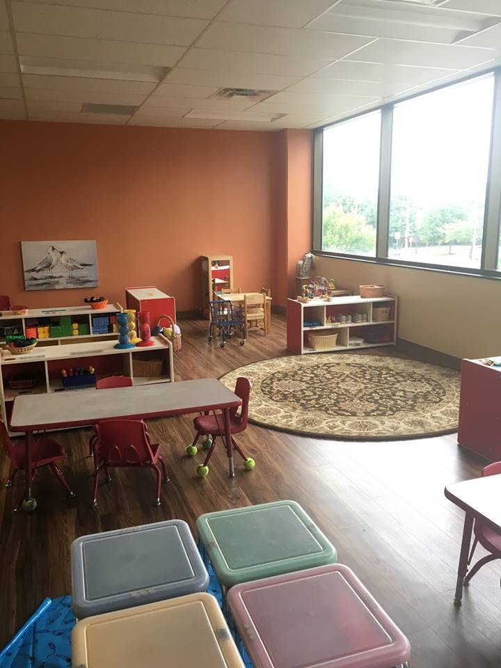 Classroom with small tables and chairs, shelves with toys, and a round rug
