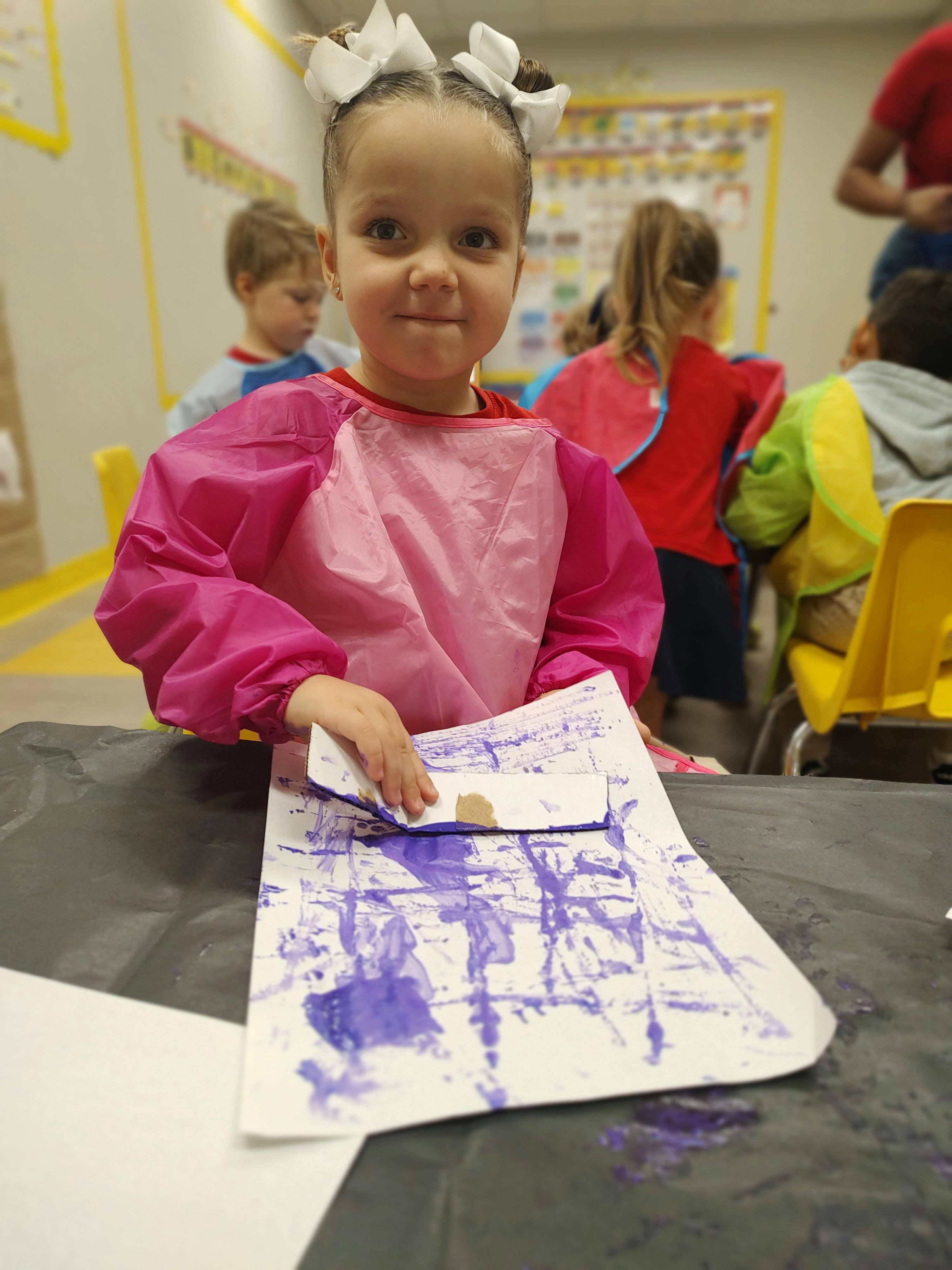 Child in a pink smock holding a paper with purple paint