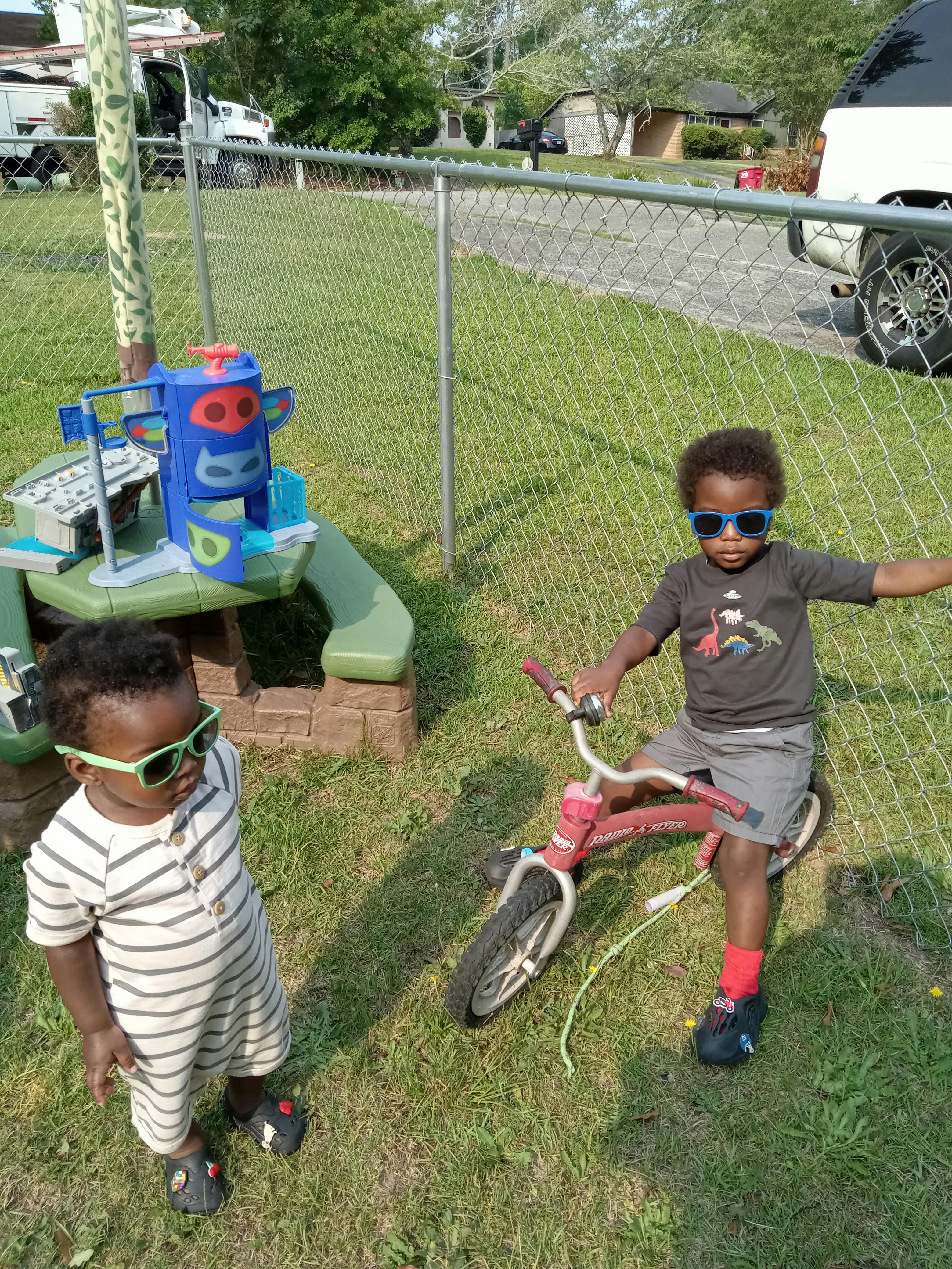 Two children wearing sunglasses in a grassy area, one on a bicycle