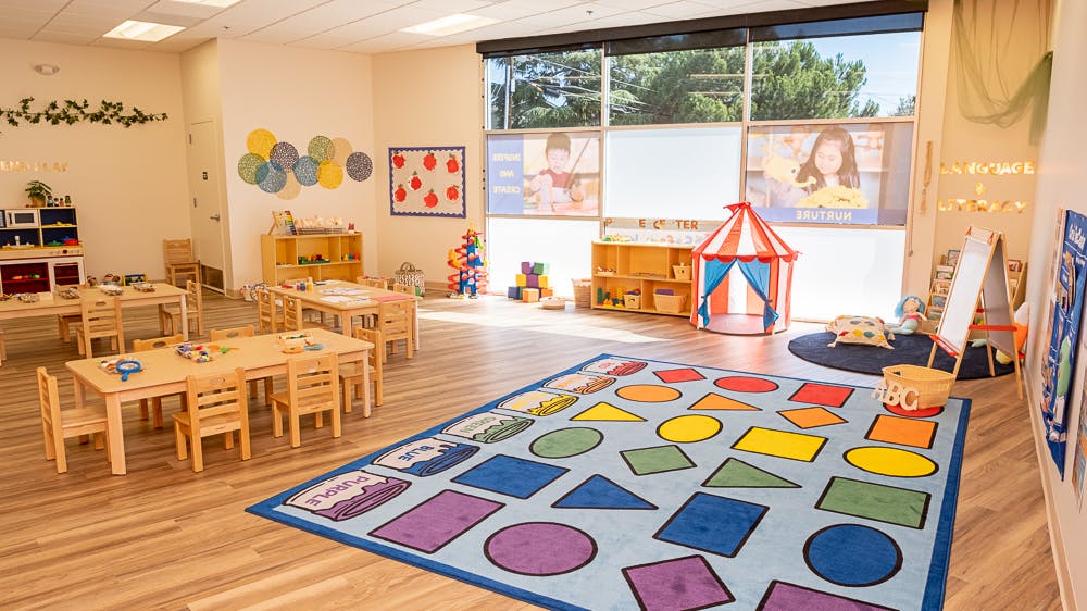 Preschool classroom with small tables and chairs, colorful rug with shapes