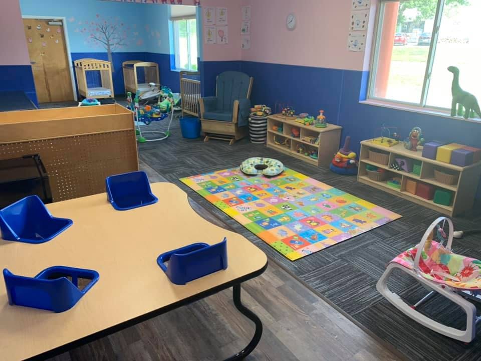 Childcare room with small table and chairs, play mat, and shelves with shelves of toys