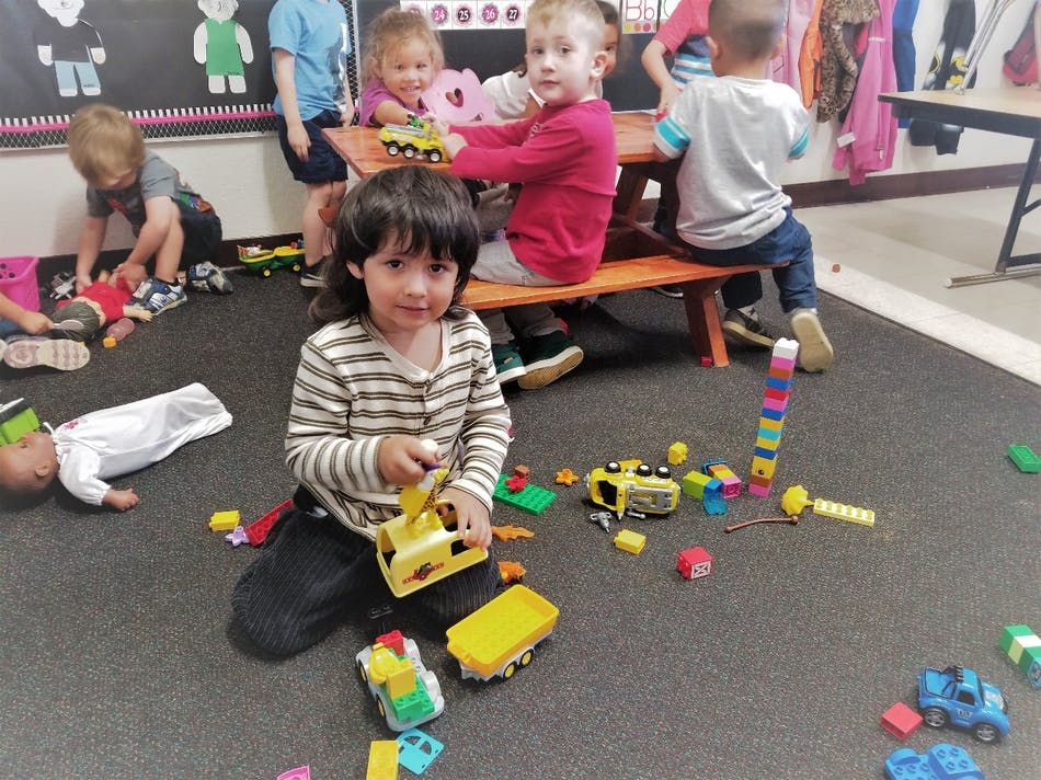 Child playing with toy truck on floor with other children and toys in a classroom